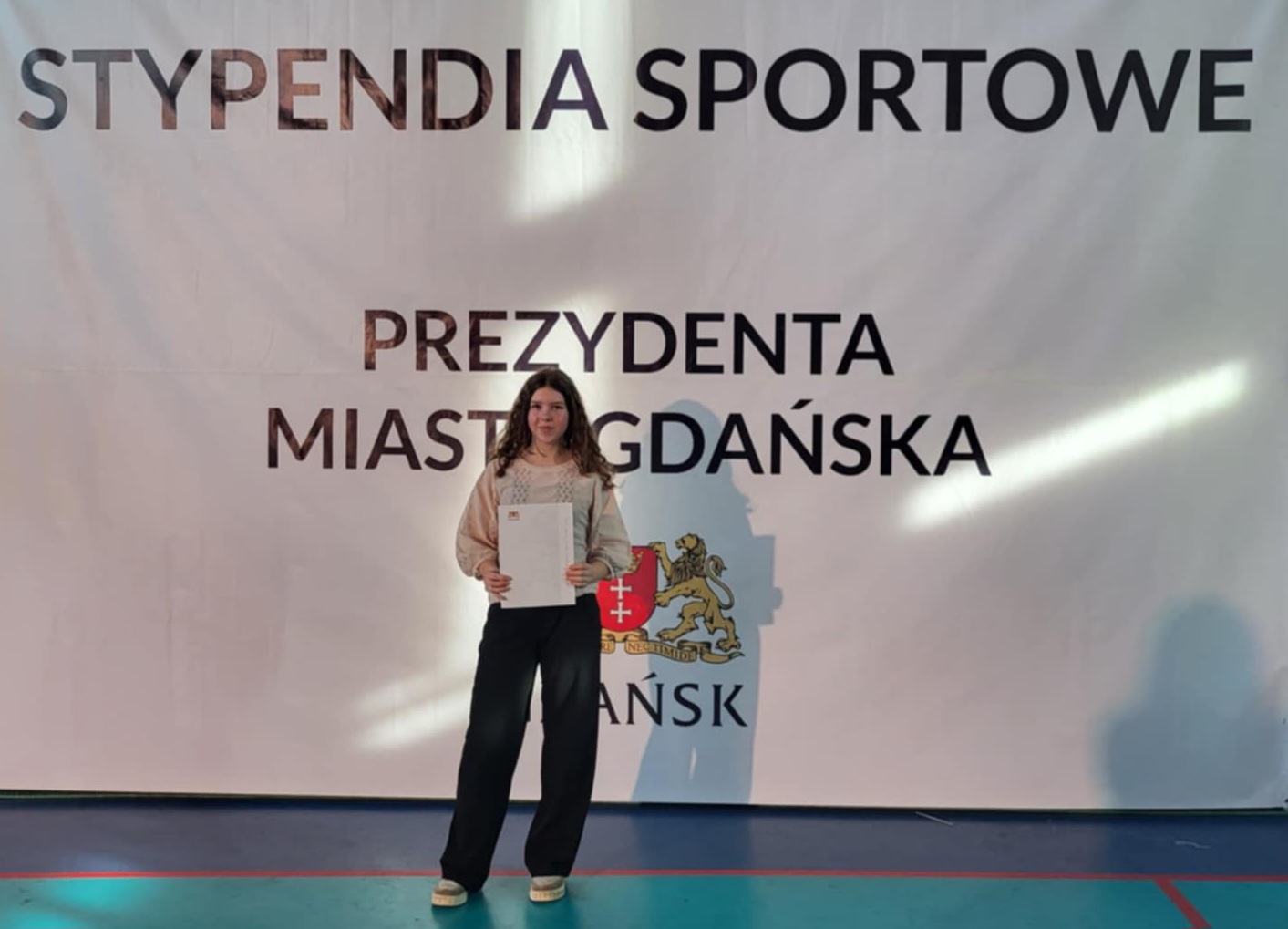 Uczennica stoi na sali sportowej na tle napisu „Stypendia Sportowe Prezydenta Miasta Gdańska”. Trzyma dyplom, ubrana w jasną bluzkę i ciemne spodnie.