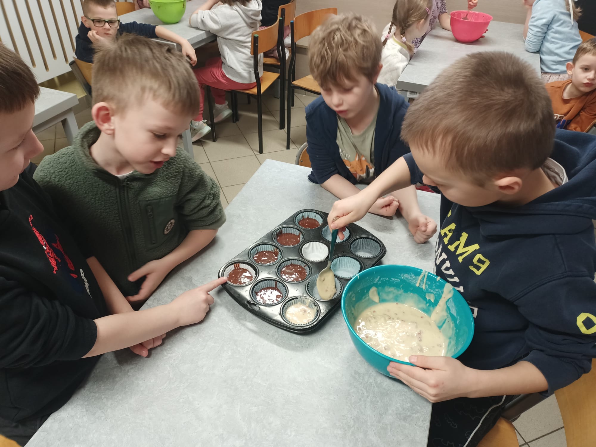 Trzech chłopców przy stoliku nakłada łyżką jasne ciasto do papilotek w formie na muffinki; obok stoi miska z ciastem.