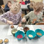 Dwoje dzieci dekoruje muffinki przy stoliku; na blacie stoją miseczki z polewą i posypkami oraz przygotowane dekoracje do ozdabiania wypieków.