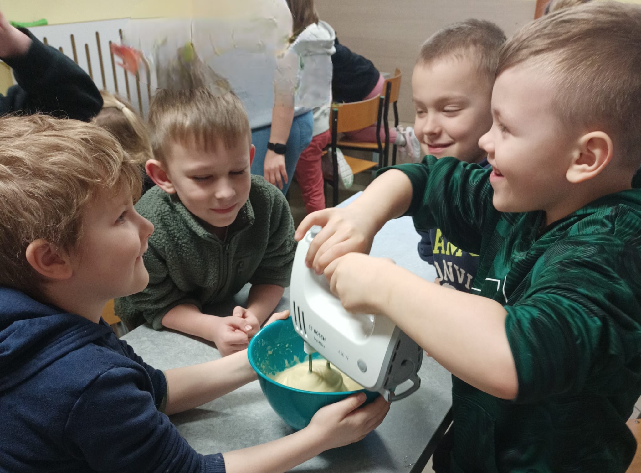 Czterech chłopców w klasie wspólnie miesza ciasto mikserem ręcznym w misce; dzieci obserwują pracę mieszadła i pomagają trzymać naczynie.