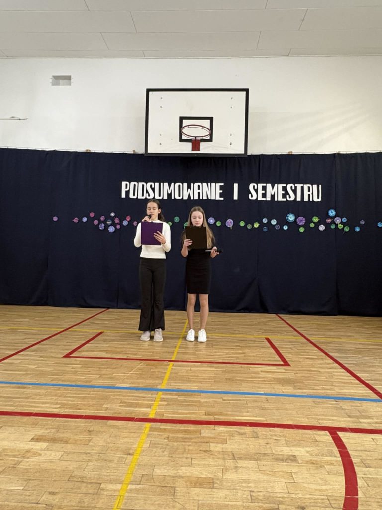 Dwie uczennice na sali gimnastycznej prowadzą apel a okazji podsumowania I semestru 2025/2026 roku