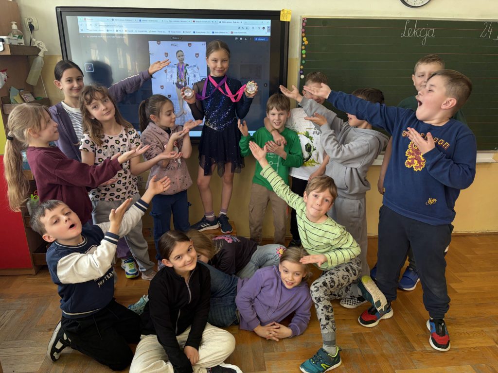Grupa trzynastu uśmiechniętych dzieci w sali lekcyjnej z drewnianą podłogą i zieloną tablicą otacza koleżankę stojącą na krześle. Dziewczynka na krześle ma na sobie ciemny strój gimnastyczny, dwa złote medale na szyi i wskazuje na duży ekran monitora za nią. Na monitorze wyświetlane jest zdjęcie tej samej dziewczynki na podium z dyplomami. Pozostałe dzieci wykonują radosne gesty podziwu, wskazują rękami na medalistkę lub klaszczą.