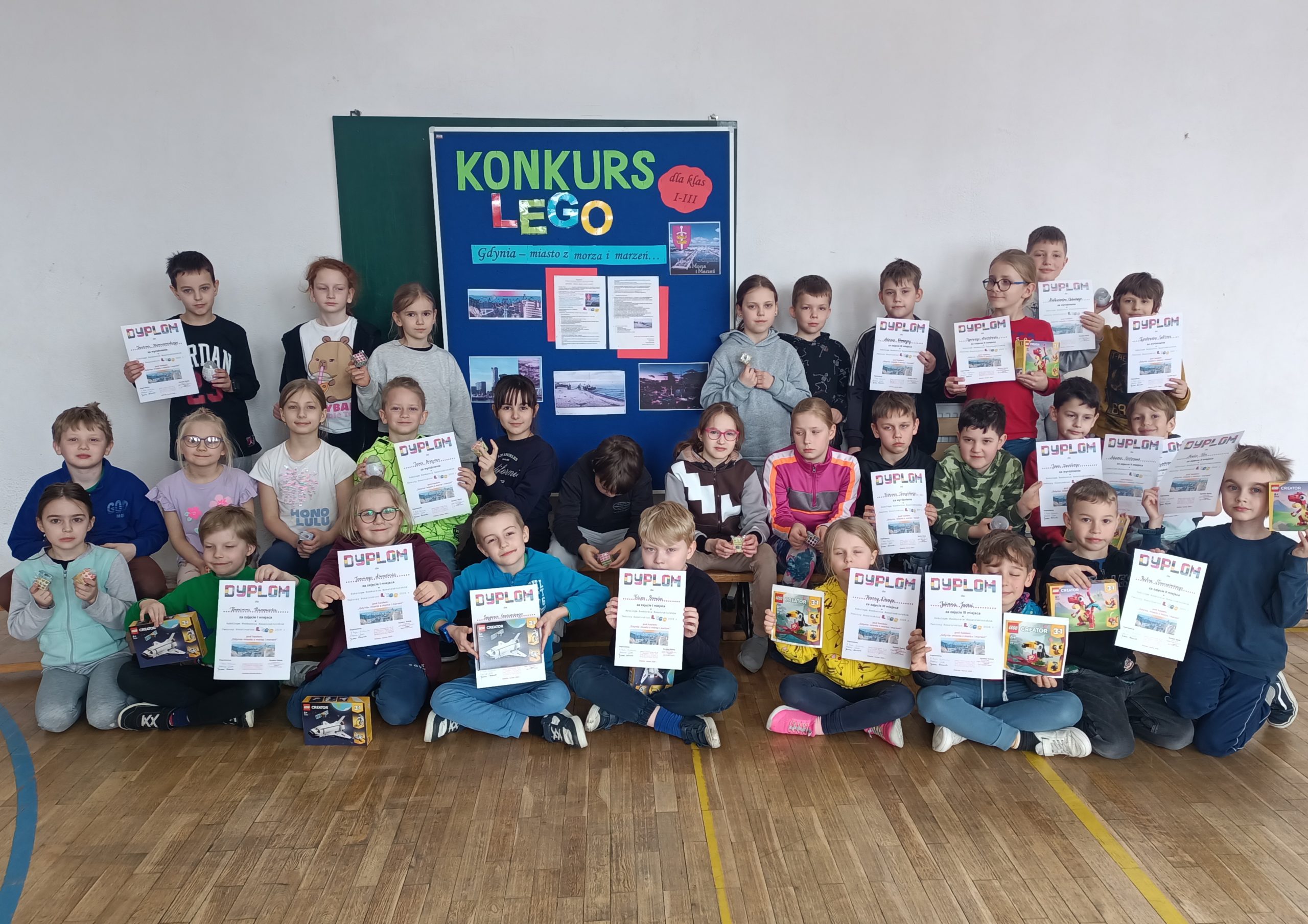 W sali gimnastycznej na tle tablicy ze napisem KONKURS LEGO stoją lub siedzą uczniowie ustawieni w trzy rzędy. Około trzydziestoosobowa grupa uczniów trzyma w dłoniach nagrody za udział w Szkolnym Konkursie Konstruktorskim LEGO, część z nich trzyma również dyplom laureata konkursu.