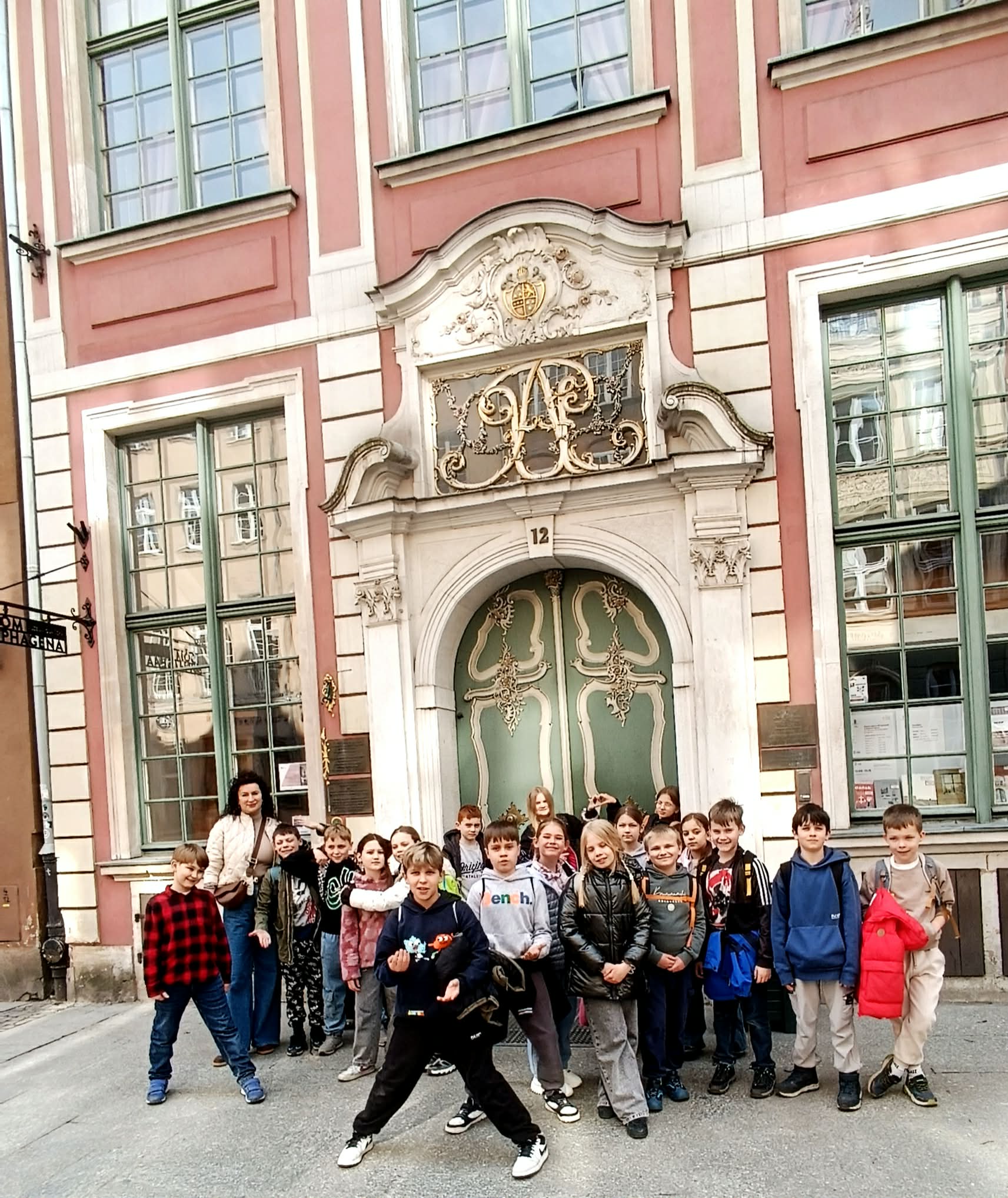 Grupa dzieci wraz z opiekunką stoi przed ozdobną, barokową kamienicą z różową fasadą i zielonymi drzwiami.