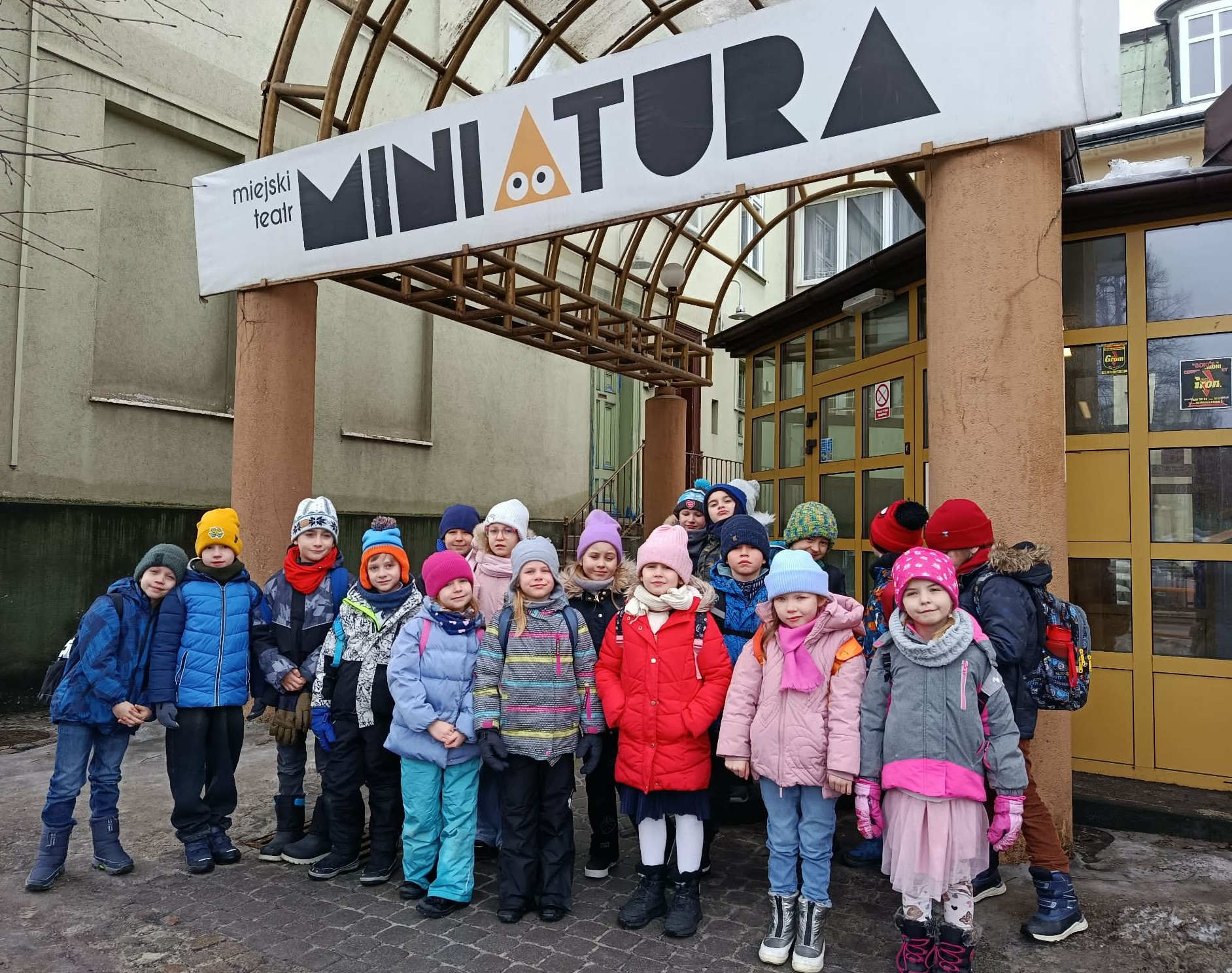 Przed budynkiem teatru stoi 20 osobowa grupa uczniów klasy 1c ustawiona w dwa rzędy. Dzieci stoją pod napisem Miejski Teatr Miniatura.