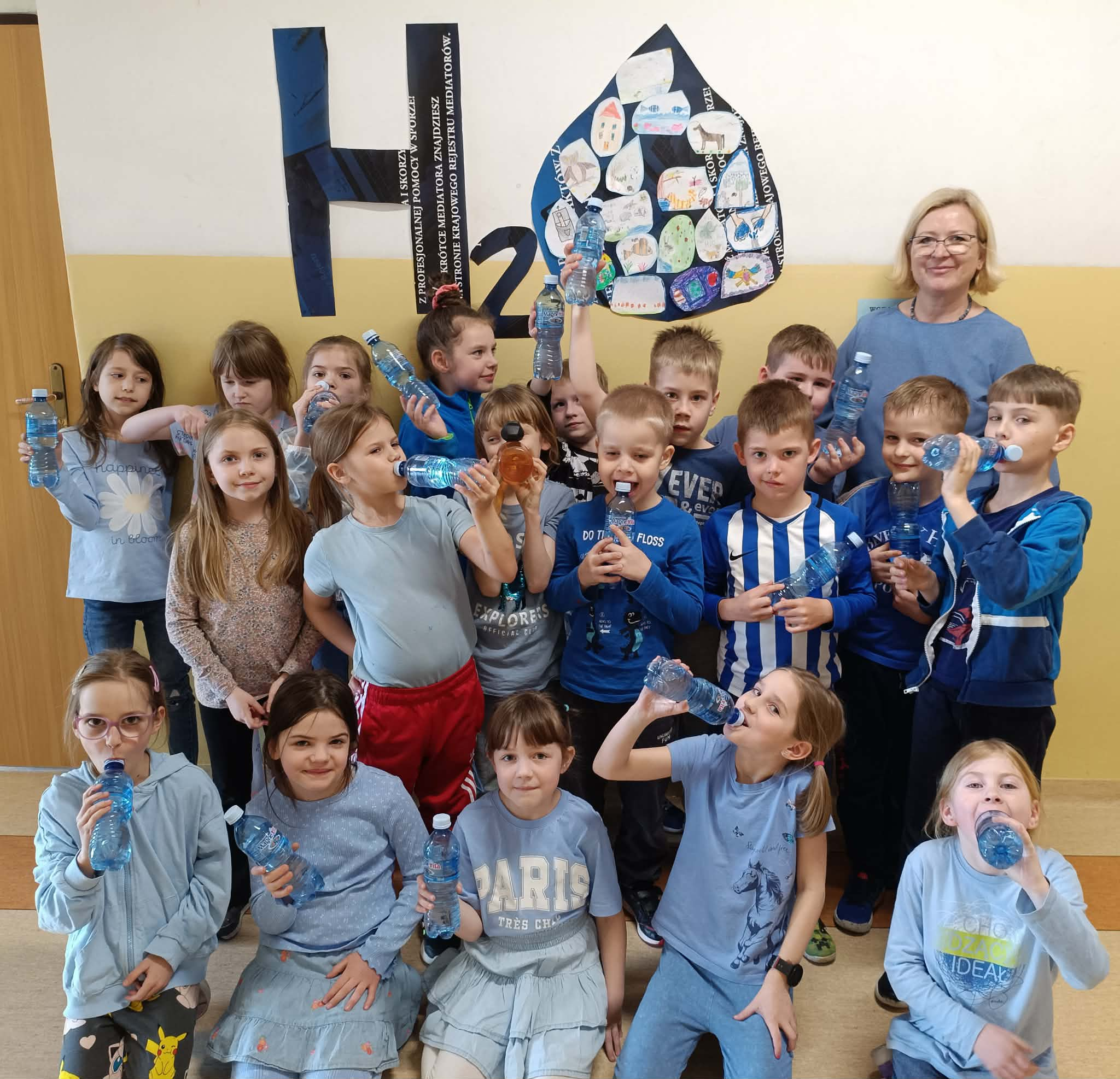 .Grupa dzieci stoi z nauczycielką na korytarzu szkolnym. Trzymają plastikowe butelki z wodą. Większość ma na sobie ubrania w odcieniach niebieskiego. Na ścianie znajduje się duża dekoracja z napisem „H₂O”, wykonana z ciemnego papieru. Obok napisu jest duża praca plastyczna w kształcie kropli wody. W środku tej kropli przyklejone są małe rysunki wykonane przez dzieci.