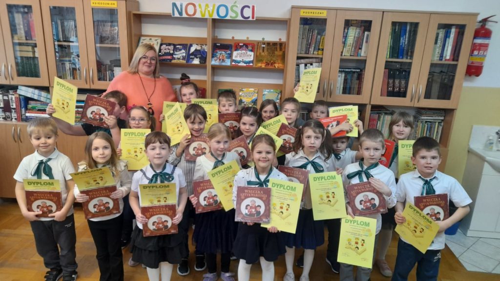 Nauczyciel bibliotekarz z grupą dziewczynek i chłopców z klasy pierwszej Na tle okna i regałów z książkami. Uczniowie i uczennice stoją, kucają lub siedzą na podłodze w bibliotece. Trzymają w rękach dyplomy z pasowania i zestawy książek do czytania.