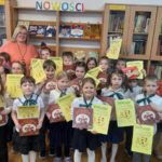 Nauczyciel bibliotekarz z grupą dziewczynek i chłopców z klasy pierwszej Na tle okna i regałów z książkami. Uczniowie i uczennice stoją, kucają lub siedzą na podłodze w bibliotece. Trzymają w rękach dyplomy z pasowania i zestawy książek do czytania.