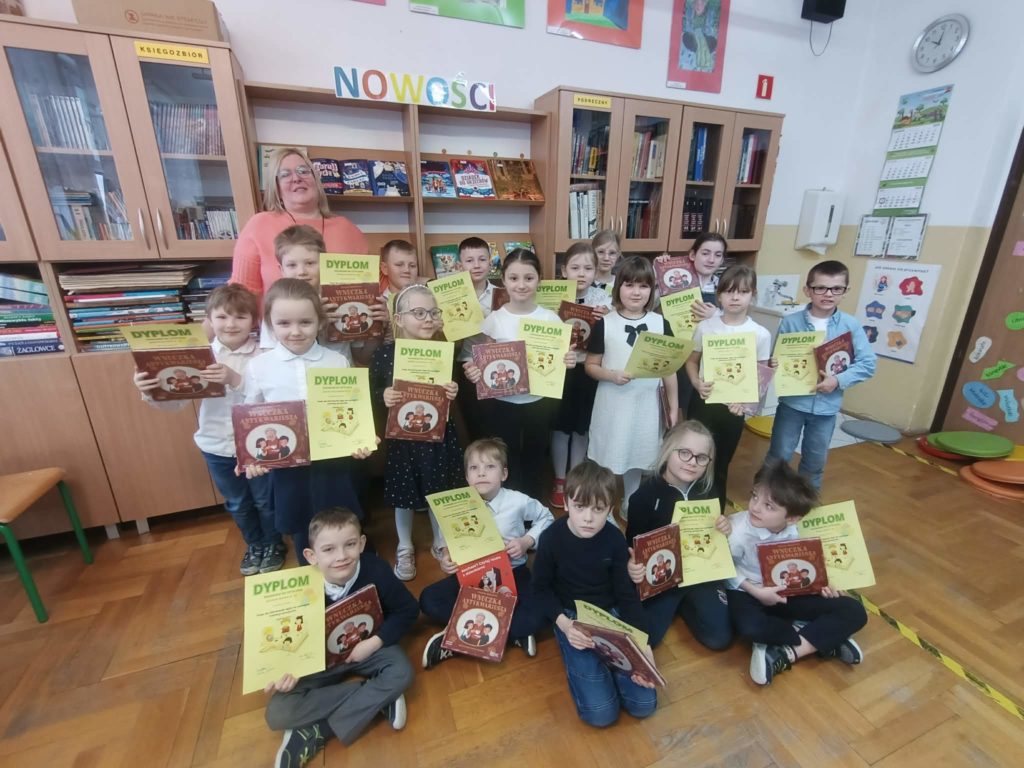 Nauczyciel bibliotekarz z grupą dziewczynek i chłopców z klasy pierwszej Na tle okna i regałów z książkami. Uczniowie i uczennice stoją, kucają lub siedzą na podłodze w bibliotece. Trzymają w rękach dyplomy z pasowania i zestawy książek do czytania.