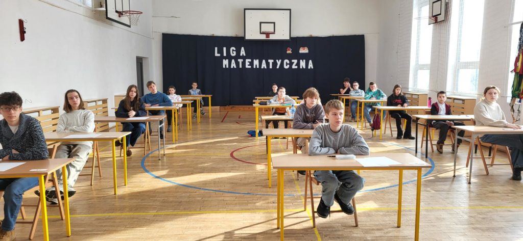 Przy kilkunastu  ławkach ustawionych w trzech rzędach na sali gimnastycznej  siedzą uczestnicy konkursu.  Z tyłu na ścianie widać napis Liga Matematyczna oraz logo szkoły 
