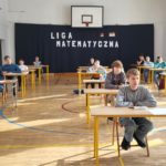 Przy kilkunastu  ławkach ustawionych w trzech rzędach na sali gimnastycznej  siedzą uczestnicy konkursu.  Z tyłu na ścianie widać napis Liga Matematyczna oraz logo szkoły 