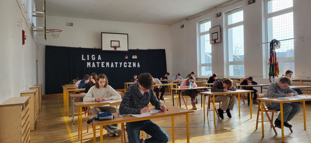 Przy kilkunastu ławkach ustawionych w trzech rzędach na sali gimnastycznej siedzą uczestnicy konkursu. Z tyłu na ścianie widać napis Liga Matematyczna oraz logo szkoły