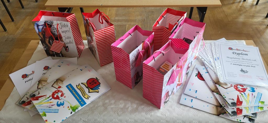 Zdjęcie wykonane z góry, przedstawiające podłużny stół przykryty białą, wzorzystą narzutą. Na stole przygotowane są nagrody konkursowe. Pośrodku stoi pięć papierowych toreb upominkowych. Trzy centralne są różowe w białe kropki, dwie skrajne są czerwono-białe w paski. Wszystkie mają ozdobne grafiki (m.in. rowerzystę, kwiaty). Po lewej i prawej stronie toreb leżą ułożone w stosy dyplomy i podziękowania. Są to zadrukowane kartki A4, niektóre w koszulkach foliowych, inne spięte. Na wierzchu stosów widoczne są dyplomy z logiem SP 47 i kolorową grafiką z przyborami szkolnymi. Wyraźnie widać nagłówek "Dyplom" i imię "Magdalena Michalczuk". Tło za stołem jest rozmyte, widać podłogę i fragmenty mebli, sugerując salę lekcyjną.