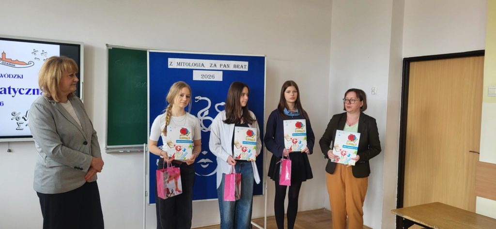 Szerokie ujęcie poziome wnętrza sali lekcyjnej. Po lewej stronie widać fragment monitora z plakatem "Liga Matematyczna". Obok niego stoi kobieta w średnim wieku z blond włosami, w szarym żakiecie i czarnych spodniach, patrząca w prawo. W centrum stoją trzy dziewczyny, na tle niebieskiej tablicy z dekoracją Meduzy. Trzymają dyplomy i różowe torby. Po prawej stronie, obok dziewczyn, stoi druga kobieta w średnim wieku z krótkimi, ciemnymi włosami, w okularach, brązowym żakiecie, jasnym topie i beżowych spodniach. Trzyma w dłoniach stos kartek, prawdopodobnie dyplomów. Sala ma jasne ściany i drewniany parkiet.