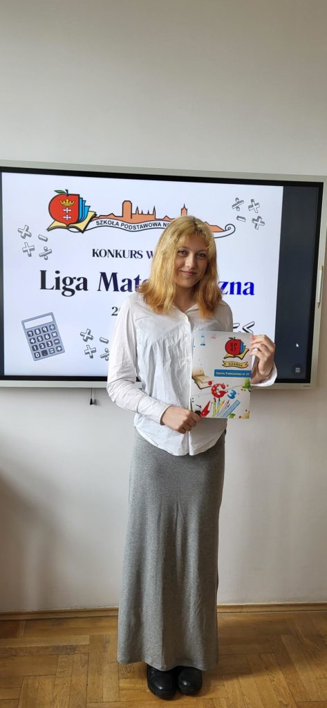 Pionowy portret młodej dziewczyny ze średniej długości blond włosami, stojącej w sali lekcyjnej. Dziewczyna ma na sobie białą koszulę z długim rękawem i bardzo długą, szarą spódnicę sięgającą do ziemi. Patrzy w stronę aparatu, lekko się uśmiechając. W obu dłoniach trzyma przed sobą zadrukowaną kartkę A4, prawdopodobnie dyplom lub podziękowanie, z kolorową grafiką szkolną. Włączony monitor z plakatem "Liga Matematyczna", fragment godła Polski na ścianie i drewniany parkiet.