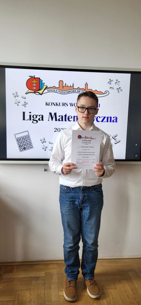 ​Pionowy portret młodego chłopaka stojącego w sali lekcyjnej. Chłopak ma krótkie, ciemne włosy, nosi okulary w czarnych oprawkach, białą koszulę z długim rękawem wpuszczoną w niebieskie jeansy oraz brązowe buty za kostkę. Stoi przodem do aparatu, patrząc w obiektyw i trzymając oburącz przed sobą zadrukowany dyplom formatu A4. Tło stanowi jasnoszara ściana, na której wisi duży monitor interaktywny. Monitor jest włączony i wyświetla plakat konkursu z logiem szkoły i napisem "Liga Matematyczna". Nad monitorem, na ścianie, wisi godło Polski (Orzeł Biały). Chłopak stoi na drewnianym parkiecie