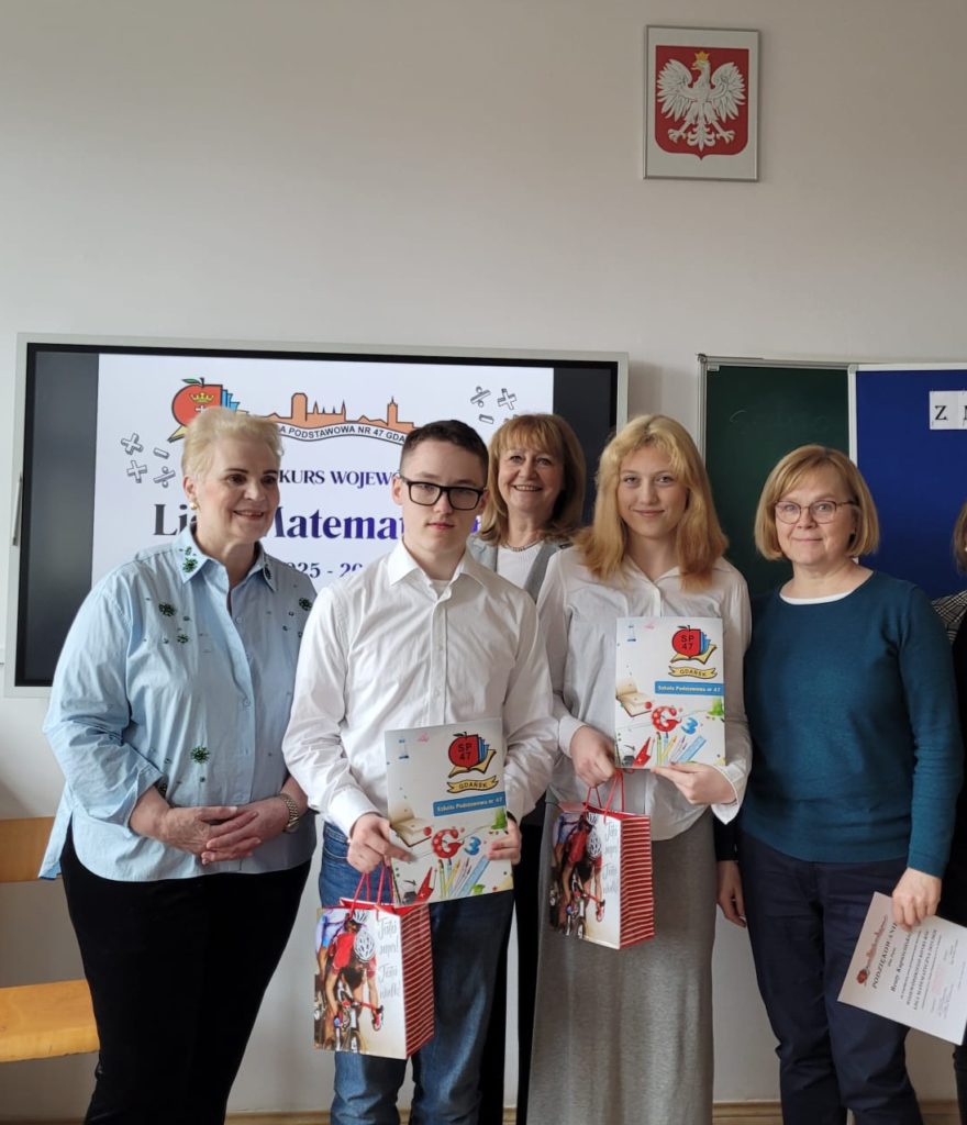 Zdjęcie grupowe sześciu osób stojących w sali lekcyjnej. Tło jest jasne, widać monitor interaktywny po lewej (z plakatem "Liga Matematyczna") i godło Polski nad nim. Od lewej stoją: Kobieta z krótkimi, jasnymi blond włosami, w błękitnej koszuli w kropki i czarnych spodniach. Chłopak w okularach i białej koszuli, trzymający dyplom i kolorową torbę (czerwono-białą). Dziewczyna z blond włosami w białej koszuli, trzymająca dyplom i kolorową torbę (czerwono-białą). Za chłopakiem widać częściowo twarz kobiety z długimi blond włosami. Kobieta po prawej od dziewczyny, w ciemnozielonym swetrze i ciemnych spodniach, w okularach. Trzyma w dłoni dyplom.