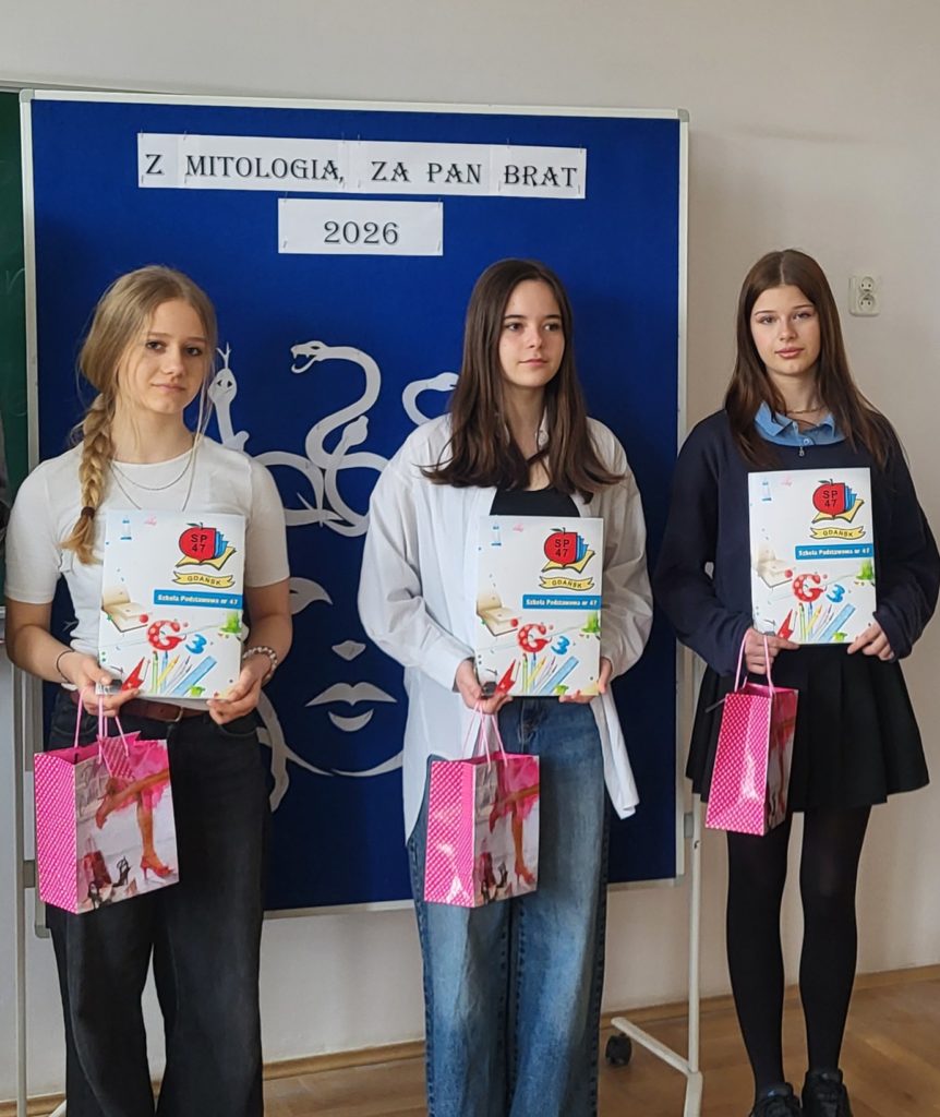 Zdjęcie grupowe trzech młodych dziewczyn stojących obok siebie w sali lekcyjnej, na tle niebieskiej tablicy z dekoracją Meduzy