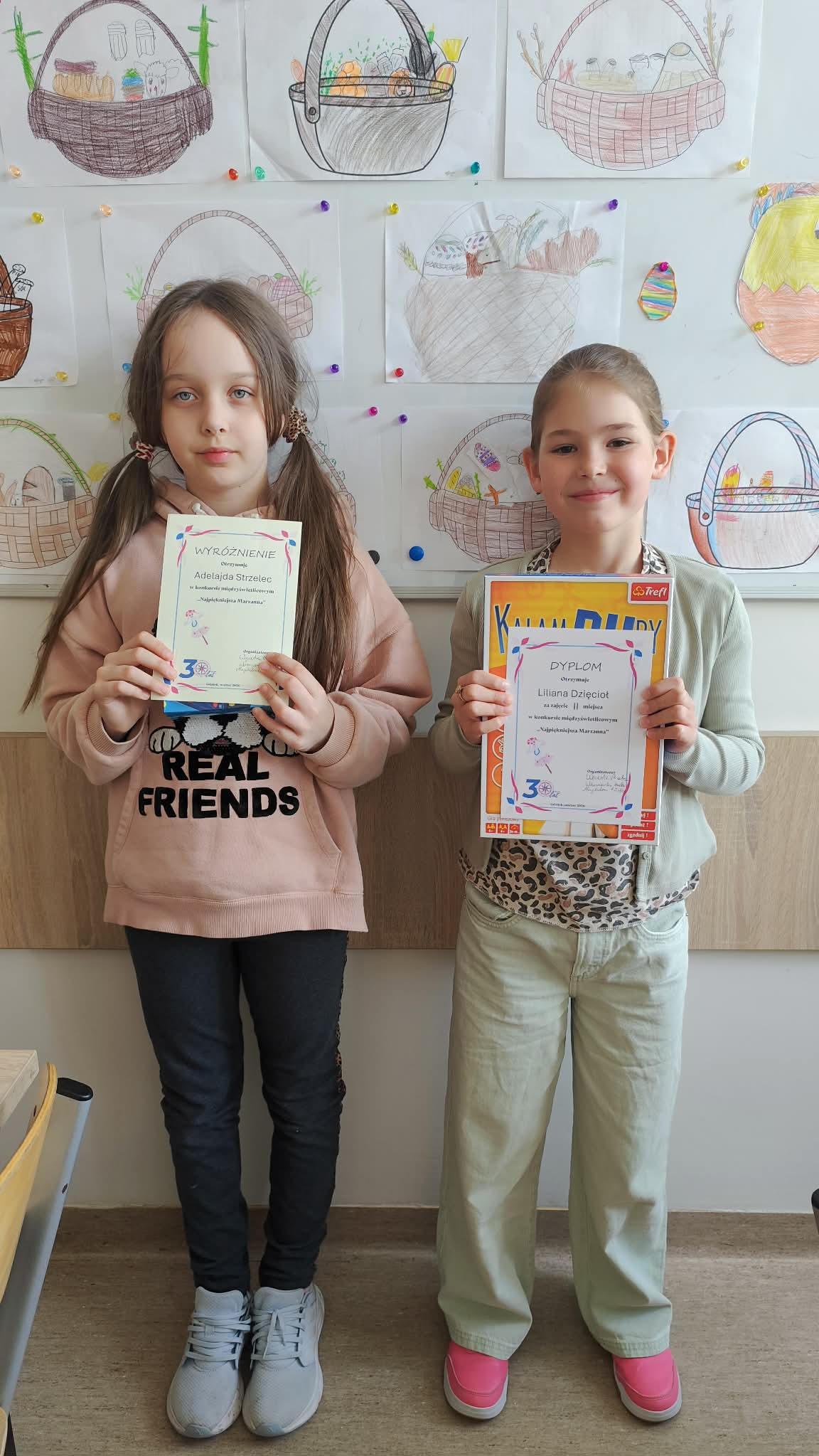 Grupa dzieci stoi na korytarzy szkolnym i trzyma dyplomy oraz gry, uśmiechając się do zdjęcia. W tle kolorowe prace plastyczne na tablicy.