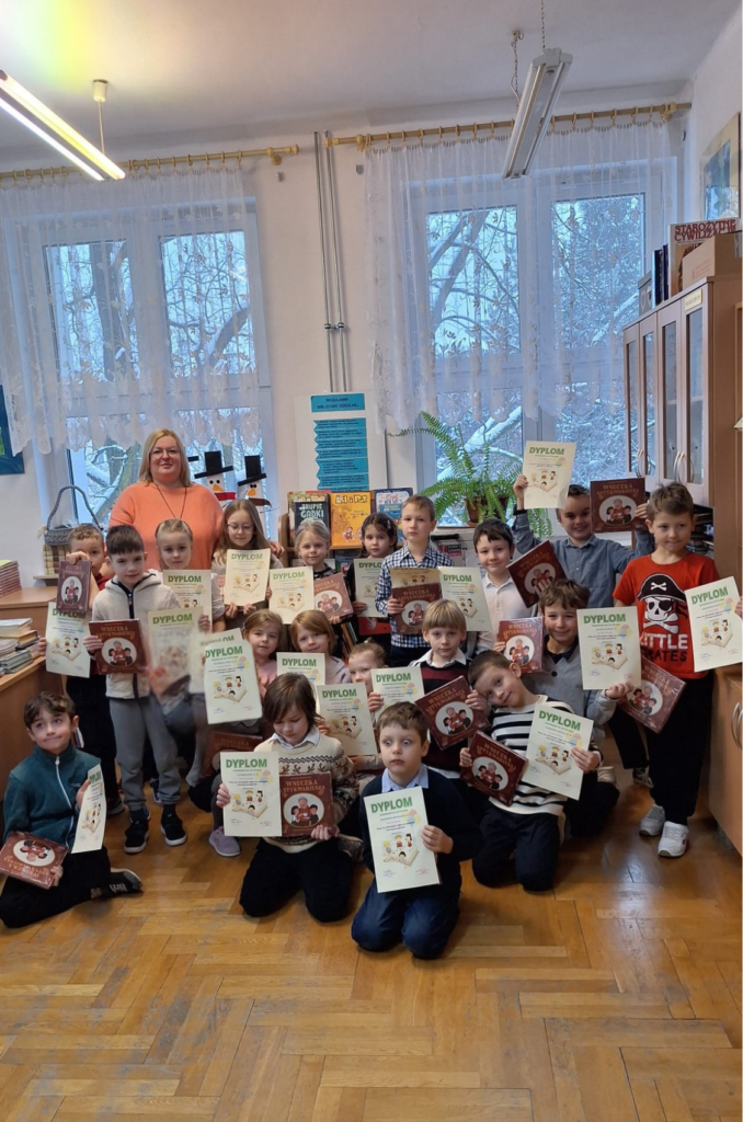 Nauczyciel bibliotekarz z grupą dziewczynek i chłopców z klasy pierwszej Na tle okna i regałów z książkami. Uczniowie i uczennice stoją, kucają lub siedzą na podłodze w bibliotece. Trzymają w rękach dyplomy z pasowania i zestawy książek do czytania.