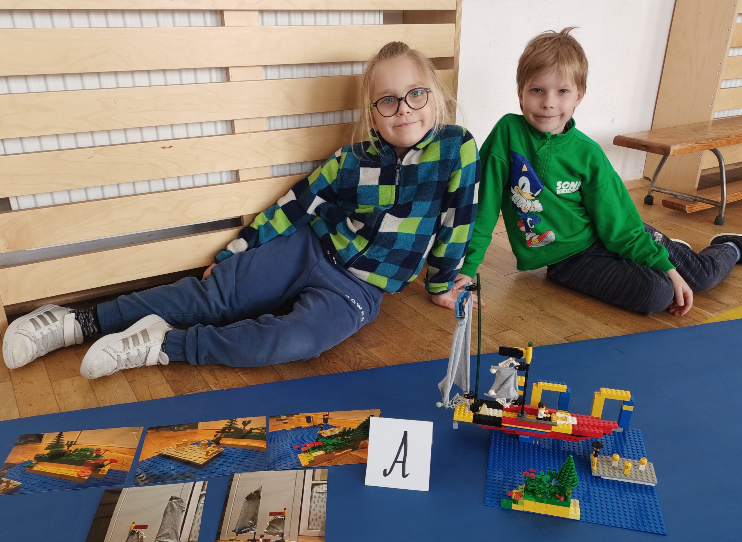 W sali gimnastycznej przed niebieskim materacem siedzi dwóch uczniów z klasy 1. Przed nimi stoi ich budowla z klocków LEGO i etykietka z literą A, obok leżą kartki z projektem budowli.