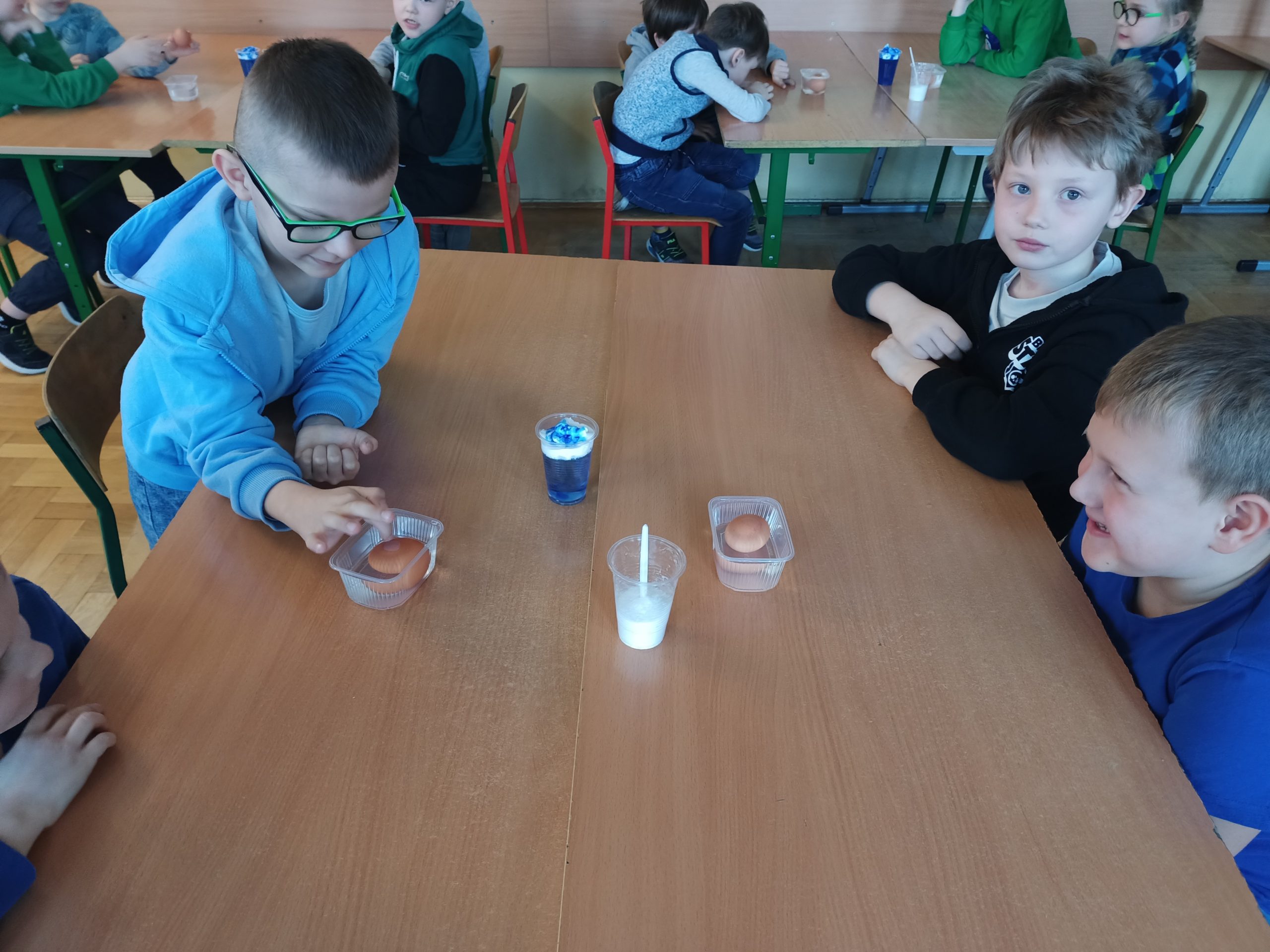 Trzech chłopców siedzi w sali lekcyjnej po obu stronach ławki. Na środku leżą dwa pojemniki z wodą i jajkami oraz dwa kubeczki z białą i niebieską cieczą. Uczniowie obserwują zjawisko wyporności.