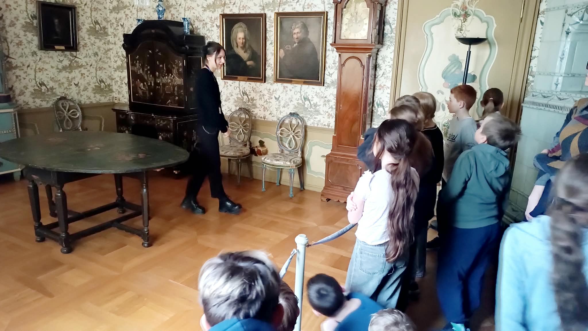 Dzieci zwiedzają historyczne wnętrze, słuchając przewodniczki w elegancko urządzonym pokoju z antycznymi meblami i zegarem.