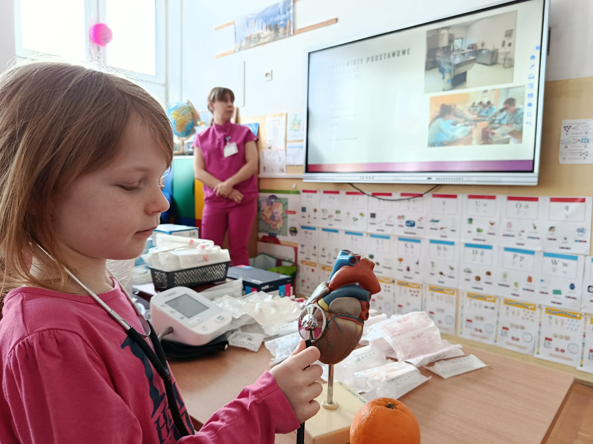 Na tle tablicy multimedialnej ze zdjęciami ze szpitala, koło stolika ze sprzętem medycznym stoi kobieta. Na pierwszym planie dziewczynka ze stetoskopem na uszach osłuchująca model serca.