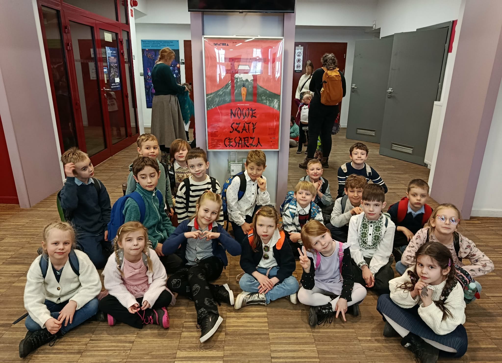 W holu Teatru Miniatura na tle plakatu z napisem ,,Nowe Szaty Cesarza" siedzą w siadzie skrzyżnym lub kucają uczniowie klasy 1c ustawieni w trzy rzędy.