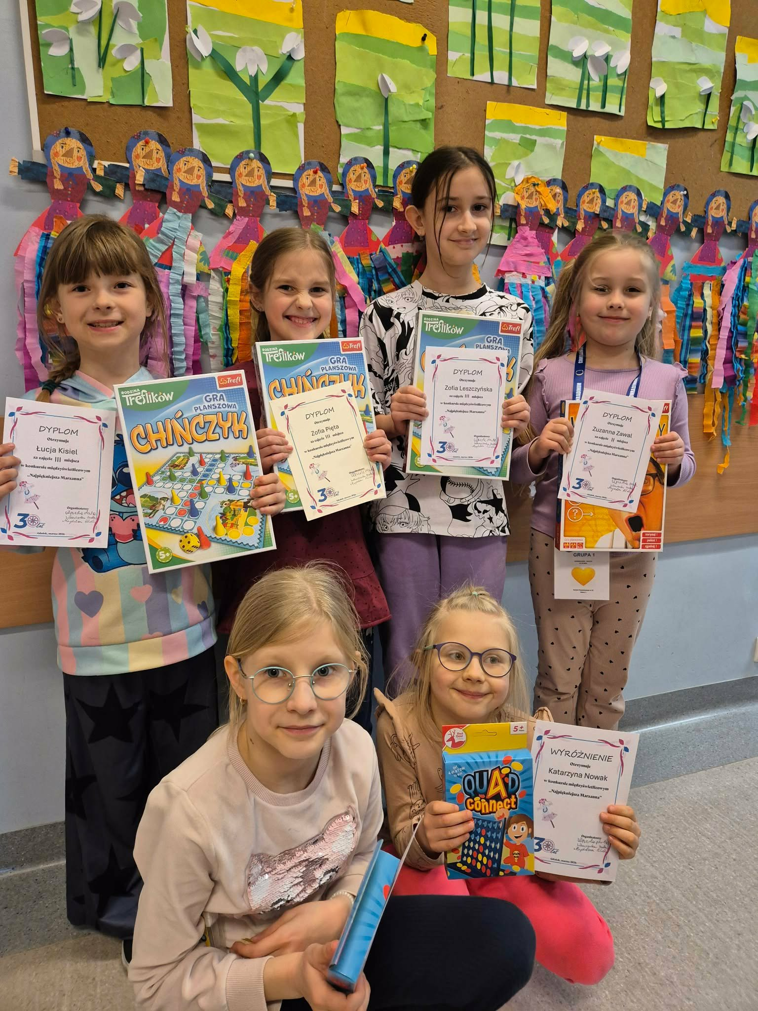 Grupa dzieci stoi na korytarzy szkolnym i trzyma dyplomy oraz gry, uśmiechając się do zdjęcia. W tle kolorowe prace plastyczne na tablicy.