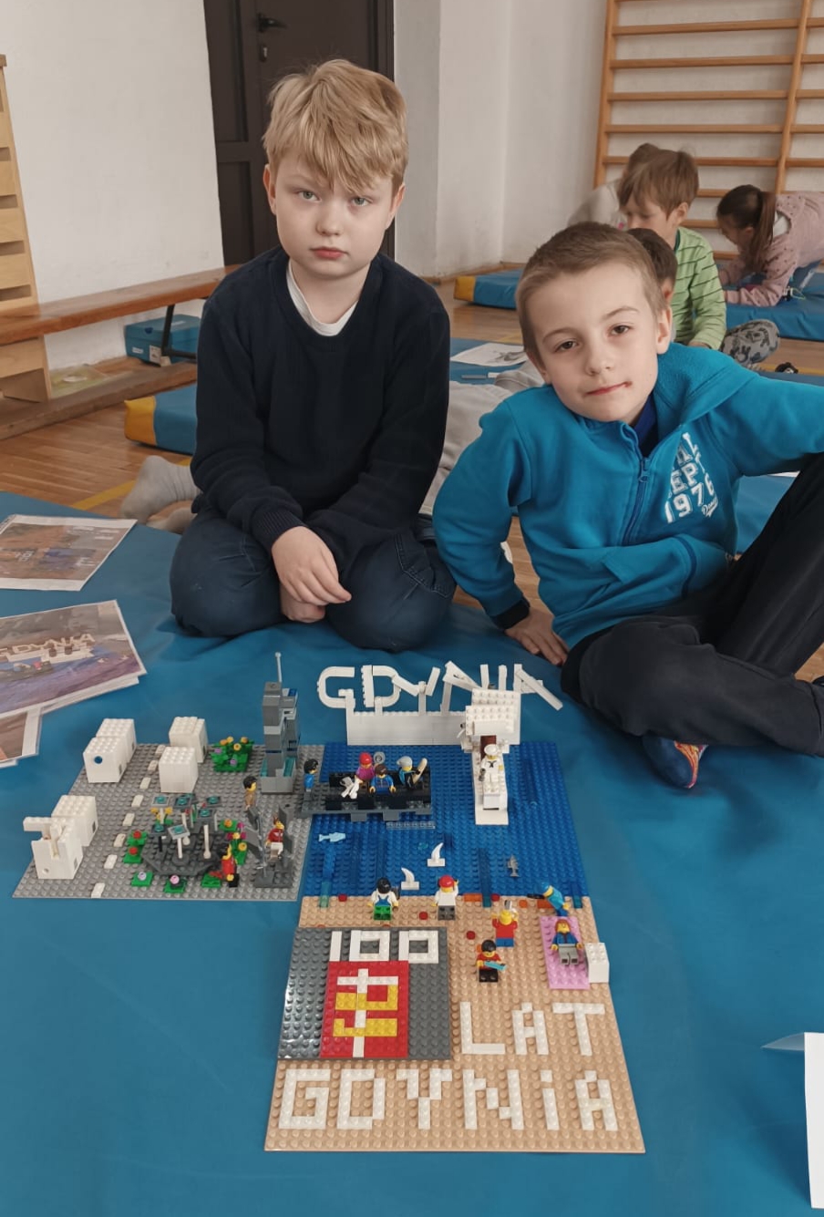 W sali gimnastycznej na niebieskim materacu siedzi dwóch uczniów z klasy 2. Przed nimi stoi ich budowla z klocków LEGO i leżą kartki z projektem.