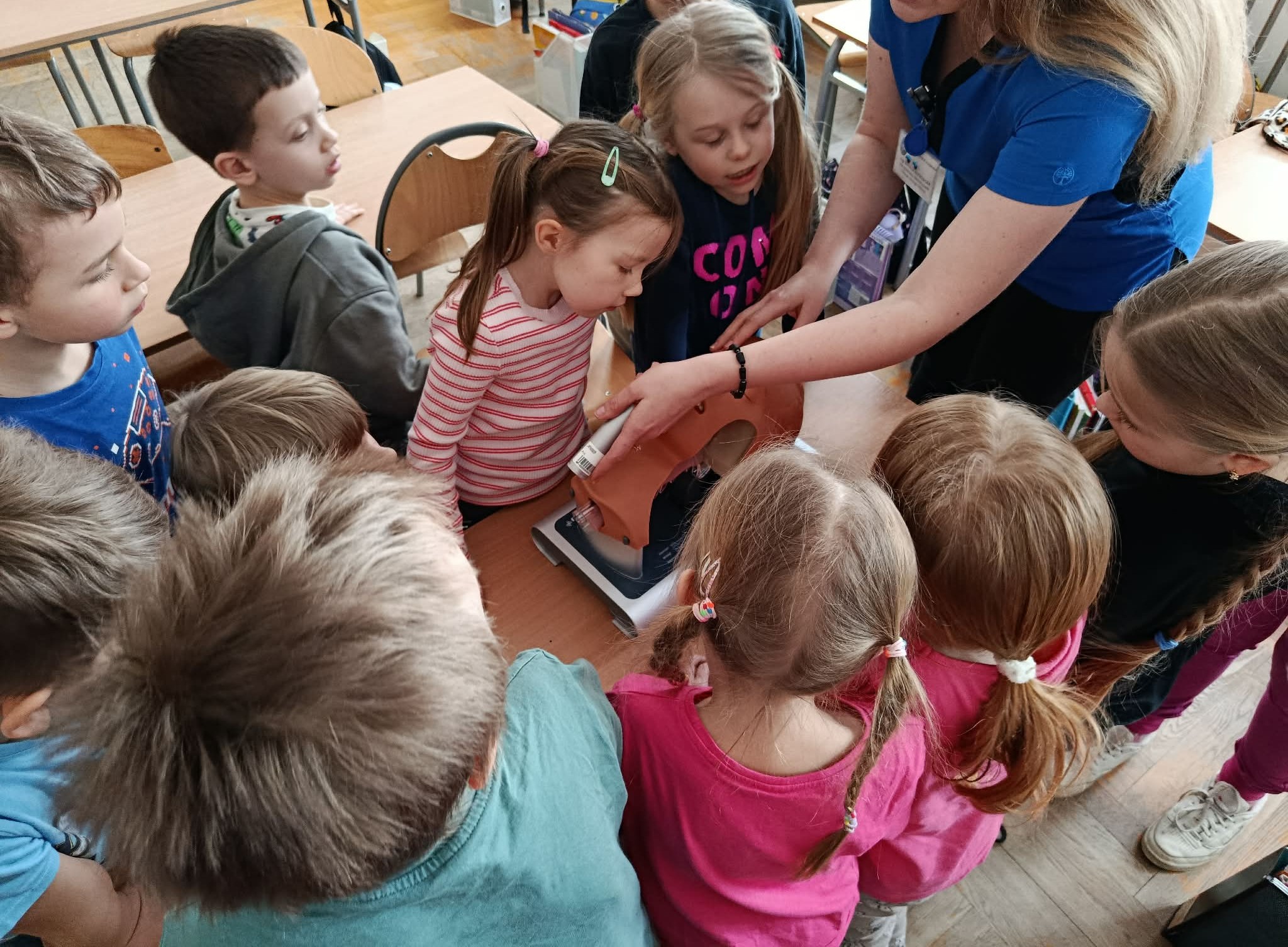 Na pierwszym planie kobieta demonstrująca udrażnianie dróg oddechowych na trenaże-rze. W koło przyglądający się uczniowie klasy 1c.