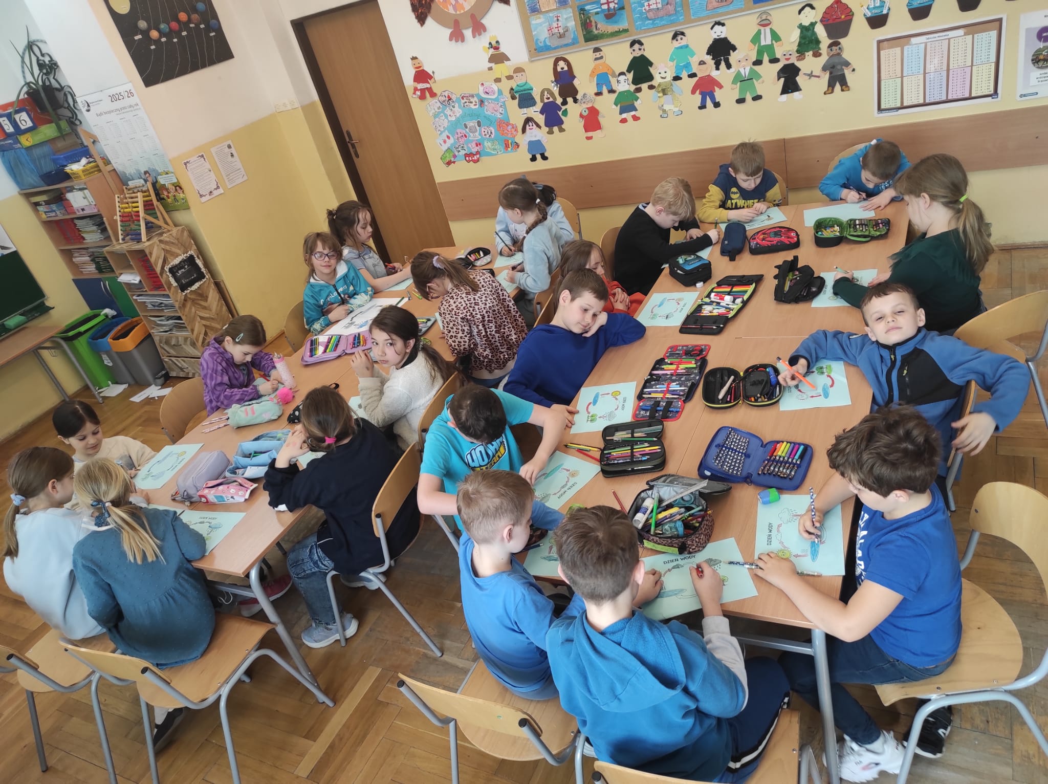  Uczniowie podzieleni na dwie grupy. Wszyscy siedzą przy stolikach, na których są kartki i piórniki. Uczniowie wykonują pracę plastyczną związaną z krążeniem wody w przyrodzie.