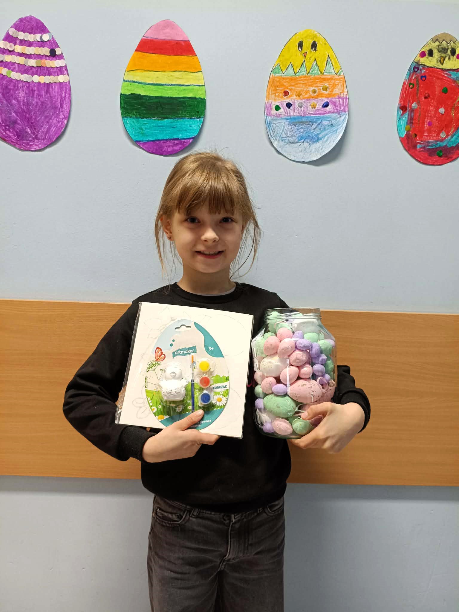 Dziewczynka trzyma słoik z kolorowymi jajkami i zestaw plastyczny na tle rysunków pisanek.