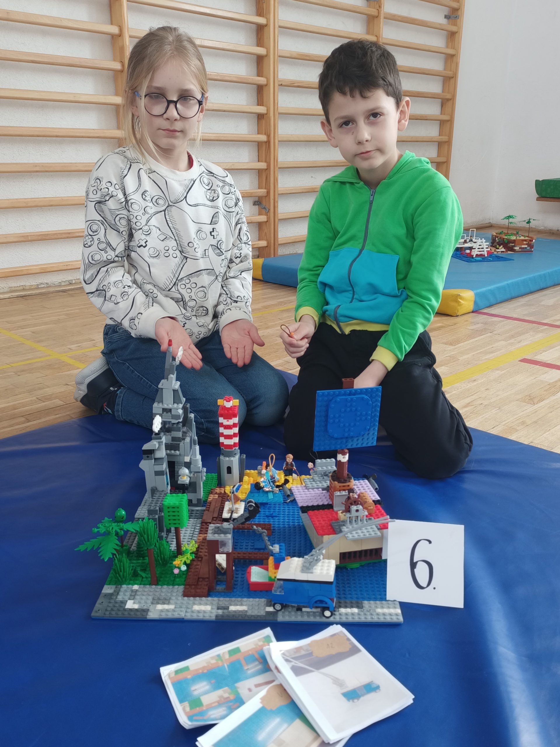 W sali gimnastycznej na niebieskim materacu siedzi dwóch uczniów z klasy 2. Przed nimi stoi ich budowla z klocków LEGO i etykietka z cyfrą 6. W tle drabinki.