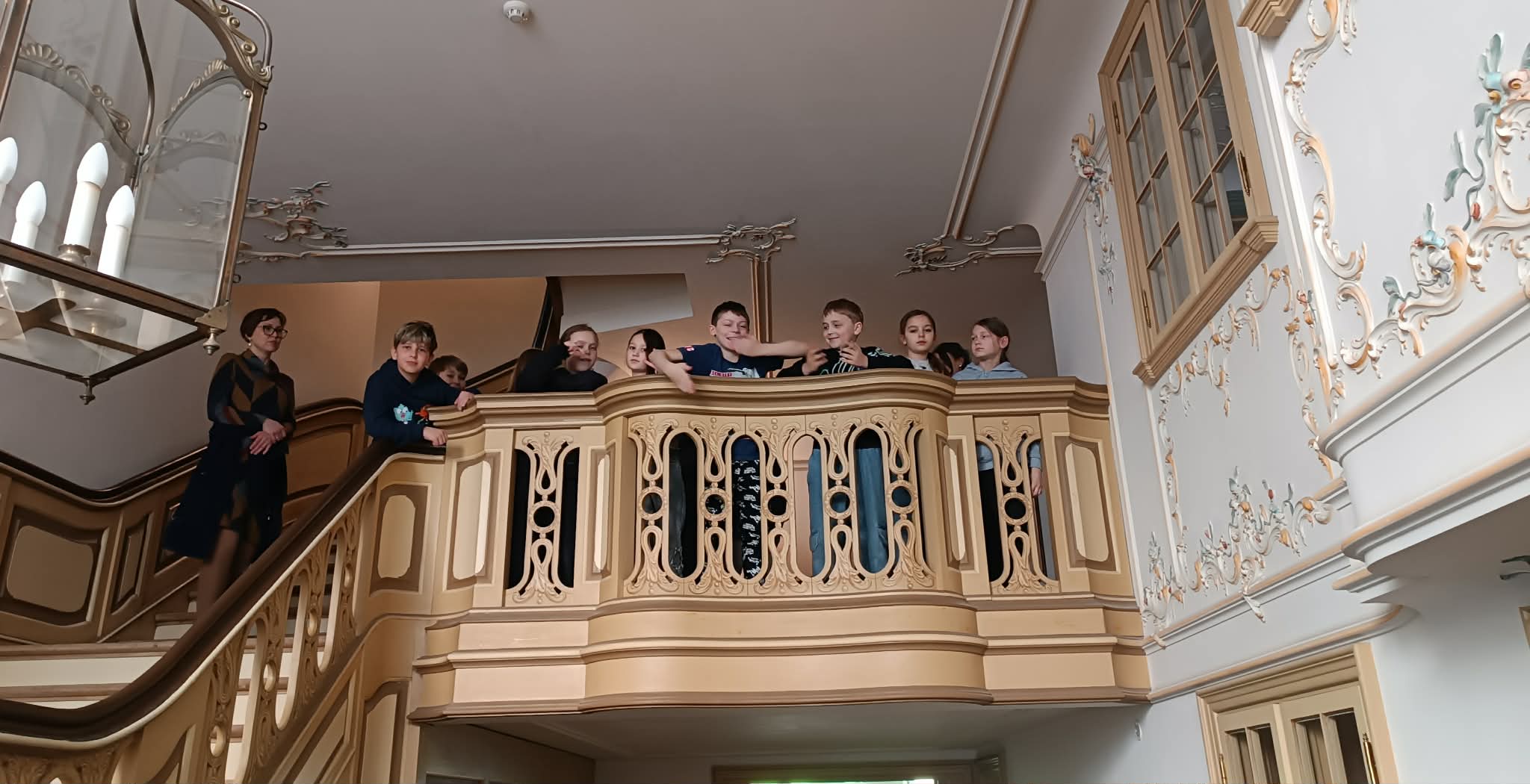 Uczniowie stoją na ozdobnym balkonie wewnątrz zabytkowego budynku i spoglądają w dół. Wnętrze charakteryzuje się eleganckimi zdobieniami i jasną kolorystyką.