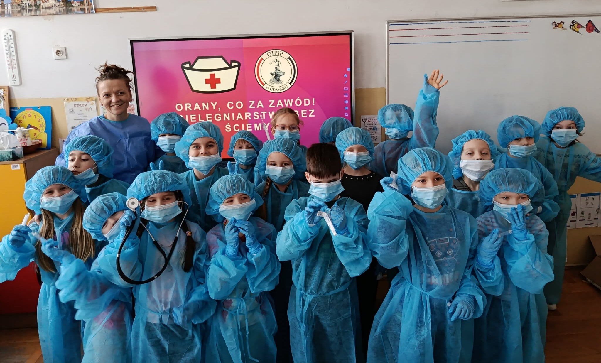 Na tle tablicy multimedialnej w napisem O RANY, CO ZA ZAWÓD! PIELĘGNIARSTWO BEZ ŚCIEMY stoją ustawieni w trzy rzędy, przebrani w szpitalny strój uczniowie klasy 1c wraz z wychowawczynią.
