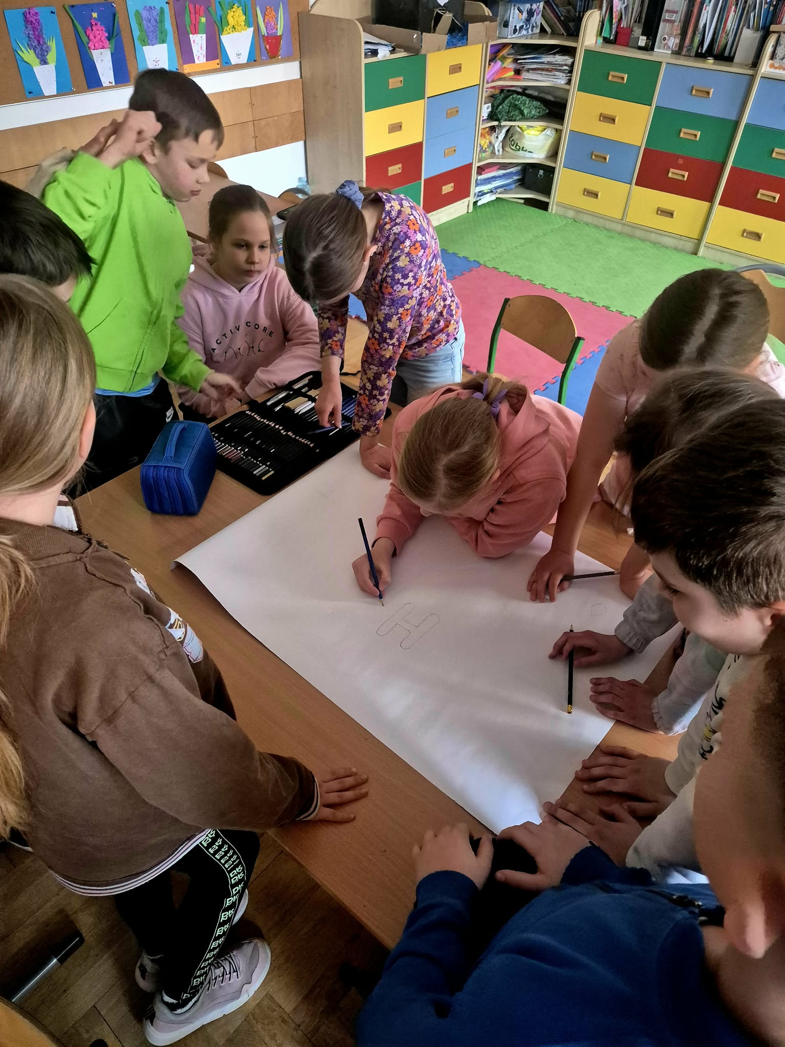 Uczniowie stoją w sali lekcyjnej wokół ławki, na której leży duży arkusz papieru. Jedna z dziewczynek szkicuje wielką kroplę wody.