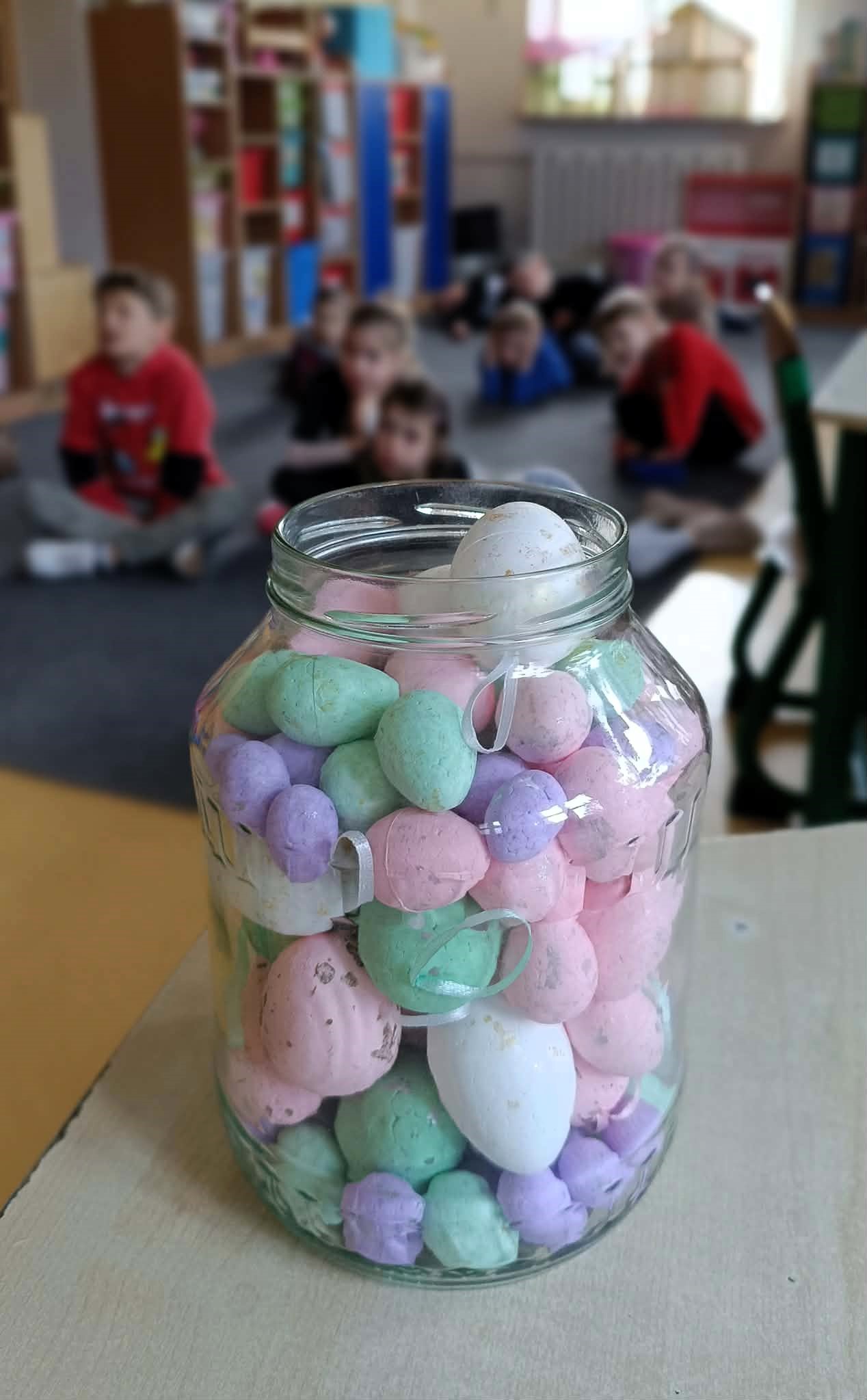 Słoik wypełniony pastelowymi jajkami, w tle rozmazane dzieci siedzące w sali.