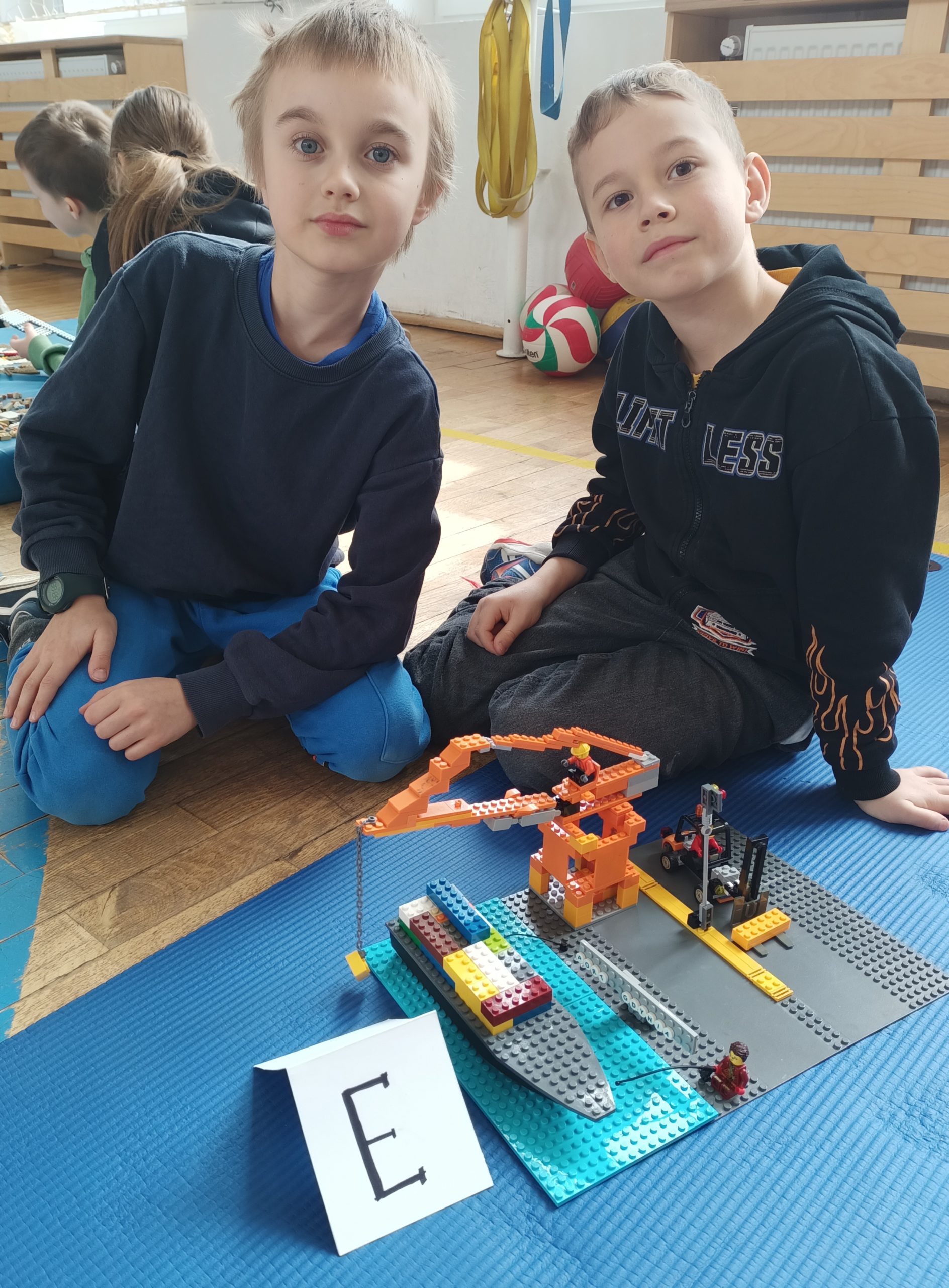 W sali gimnastycznej na niebieskim materacu siedzi dwóch uczniów z klasy 1. Przed nimi stoi ich budowla z klocków LEGO i etykietka z literą E. W tle piłki i drewniana zabudowa kaloryfera.