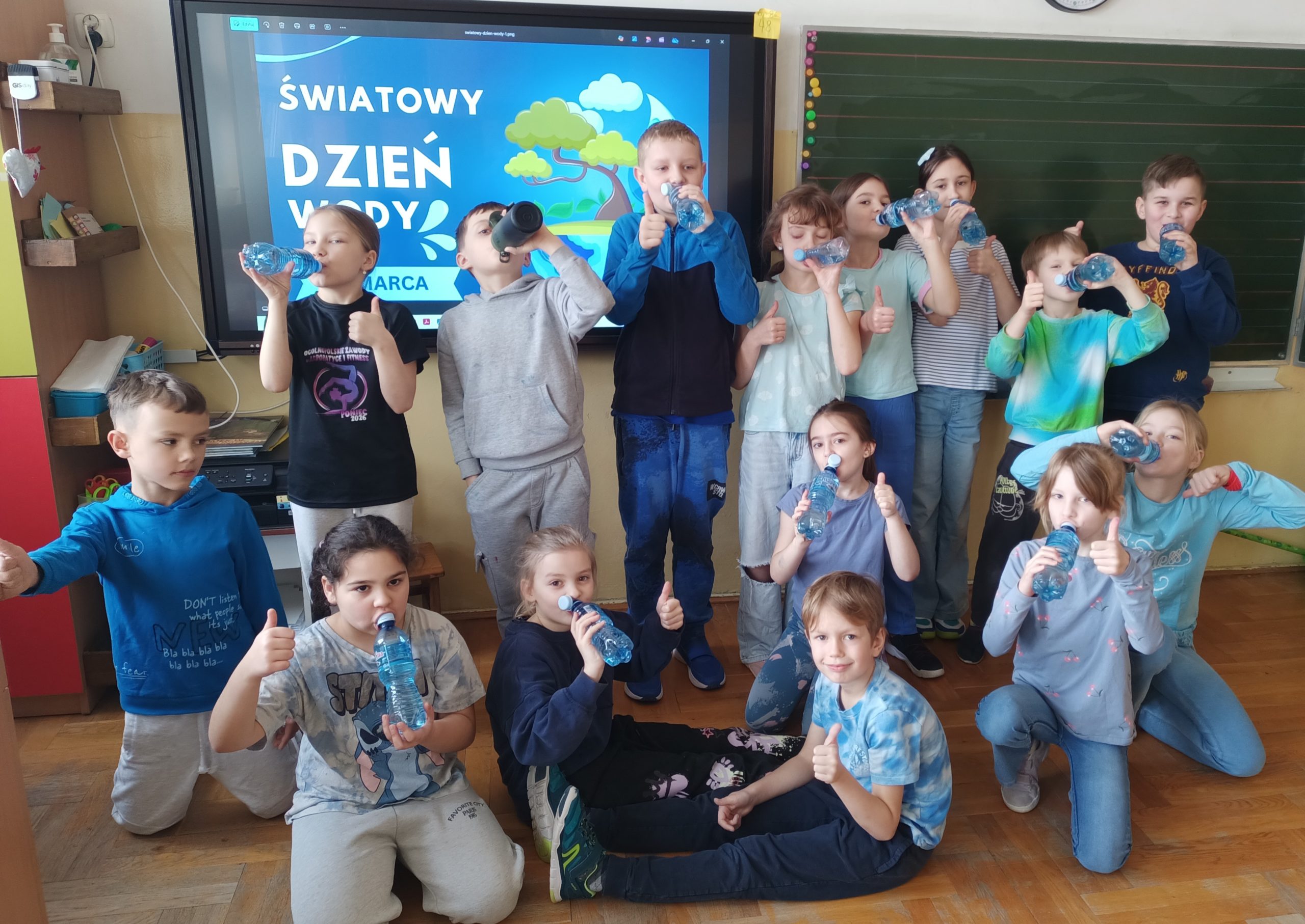  Uczniowie ustawieni w dwóch rzędach pozują na tle tablicy z napisem "Światowy Dzień Wody". Wszyscy trzymają plastikowe butelki z wodą. Większość ma na sobie ubrania w odcieniach niebieskiego.