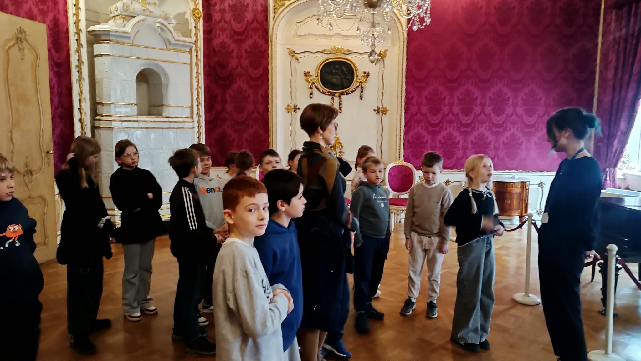  Dzieci wraz z przewodnikiem stoją w bogato zdobionej sali z czerwonymi ścianami i stylowymi meblami. Uczniowie słuchają opowieści o dawnych czasach i życiu mieszkańców