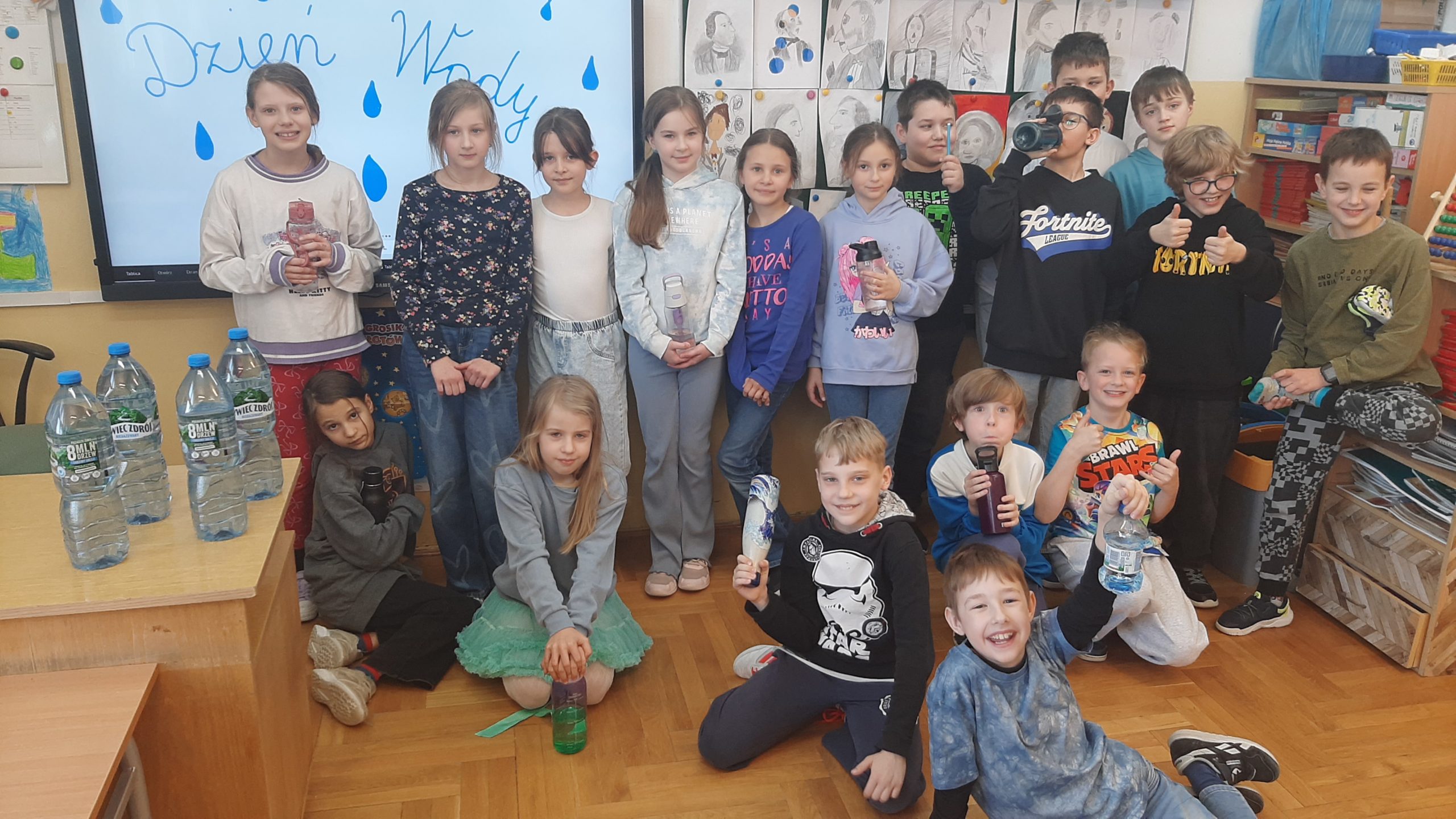 Grupa dzieci ustawiona w rzędzie pod ścianą. Przed nimi kilkoro dzieci klęczy. Za stojącymi od lewej tablica interaktywna z napisem Dzień Wody oraz tablica z portretami mężczyzn. Po lewej na biurku stoją cztery butelki wody Żywiec Zdrój. W ubiorze dzieci dominuje kolor niebieski. Niektóre dzieci trzymają w dłoniach bidony z wodą.