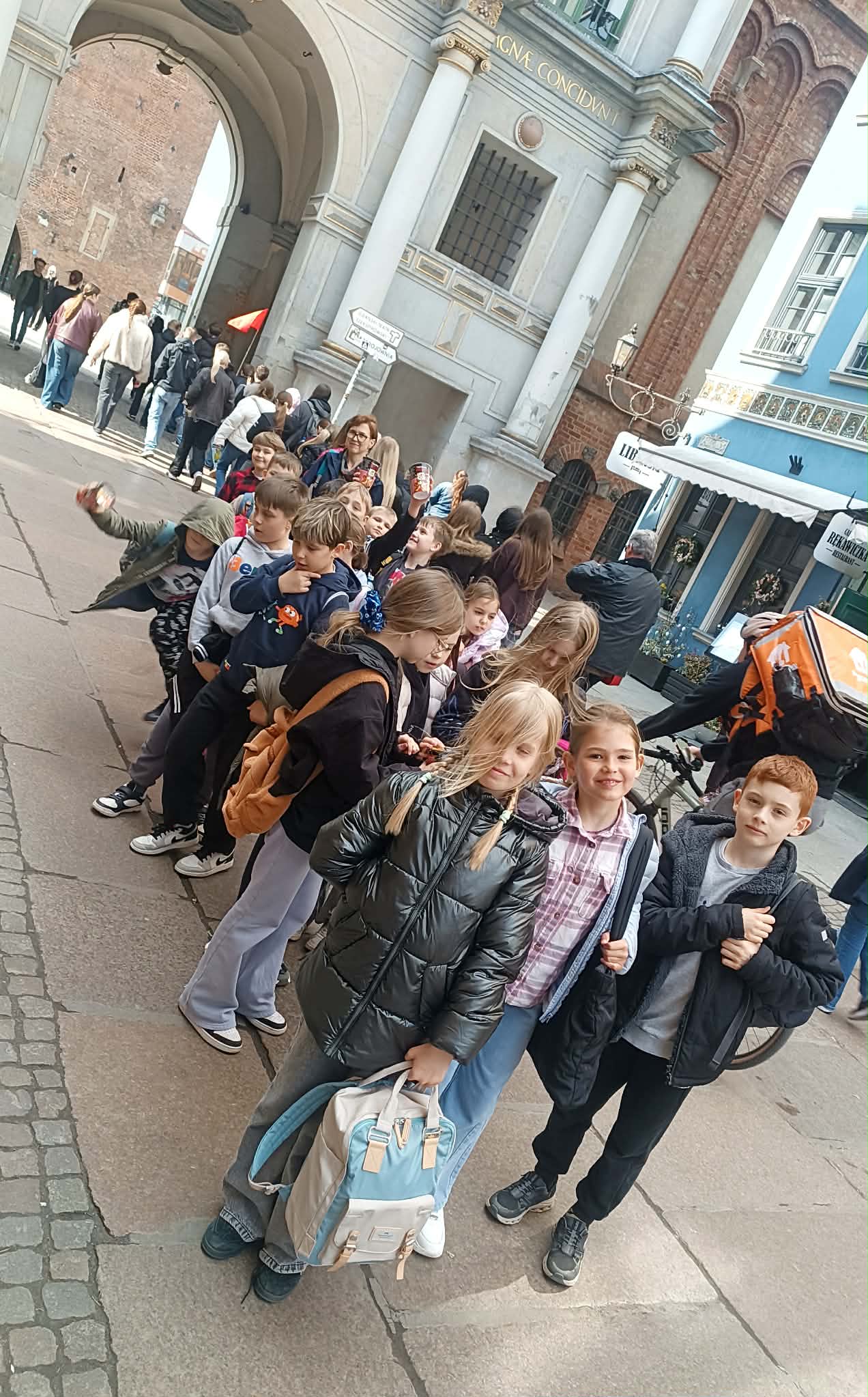Grupa uczniów klasy 3 stoi na ulicy w centrum miasta, w pobliżu zabytkowej bramy. Dzieci są uśmiechnięte, ubrane w kurtki i plecaki, przygotowane do zwiedzania.