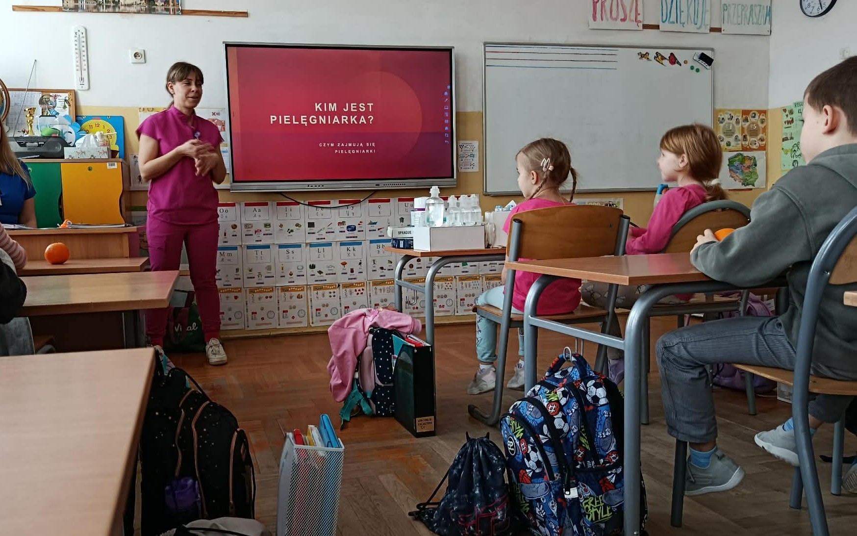 Na tle tablicy multimedialnej z napisem KIM JEST PIELĘGNIARKA? Stoi kobieta prze-brana w strój pielęgniarki. Przed nią stolik ze sprzętem medycznym i uczniowie w ławkach.