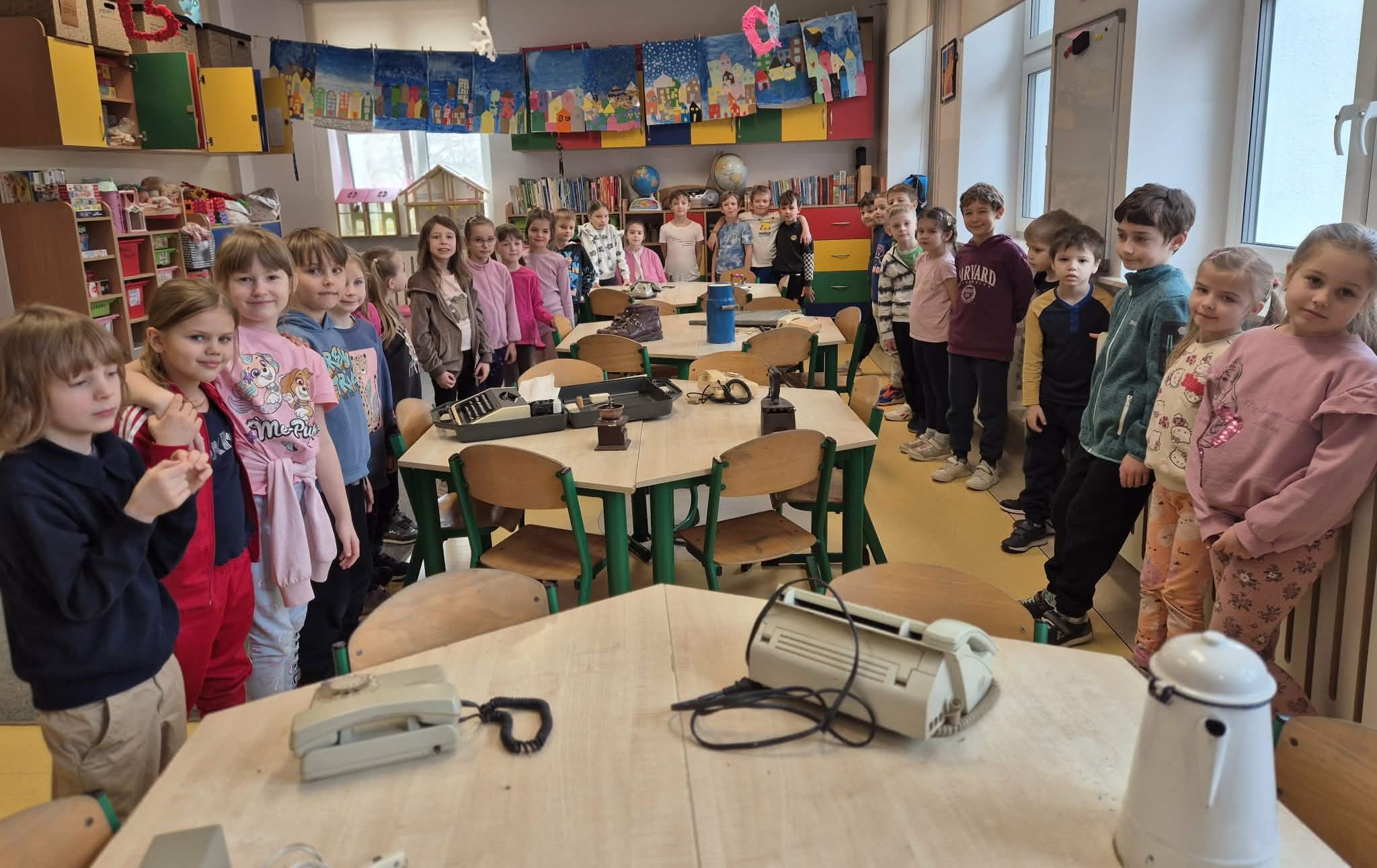 Grupa dzieci stoi w sali wokół zestawionych stołów. Na blatach leżą stare urządzenia, m.in. telefon stacjonarny i inne sprzęty elektroniczne. W tle widać kolorowe szafki, półki z książkami oraz prace plastyczne przedstawiające zimowe miasteczka zawieszone nad oknami.