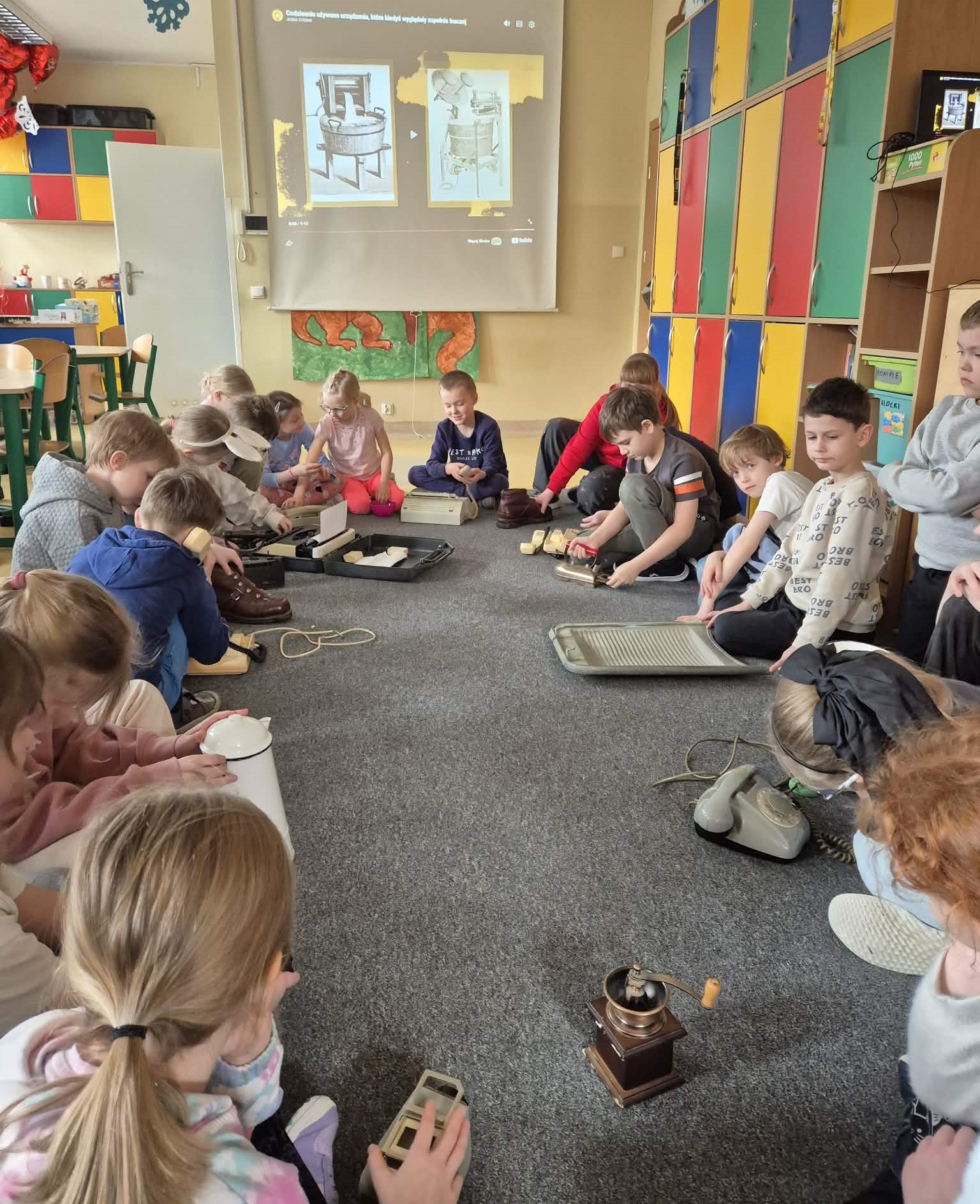 Dzieci siedzą w kręgu na dywanie w sali i oglądają stare przedmioty codziennego użytku, m.in. telefon z tarczą, ręczny młynek do kawy, tarkę do prania, żelazko i dzbanek. Na ścianie za nimi wyświetlana jest prezentacja z ilustracjami dawnych urządzeń, a obok stoją kolorowe szafki.