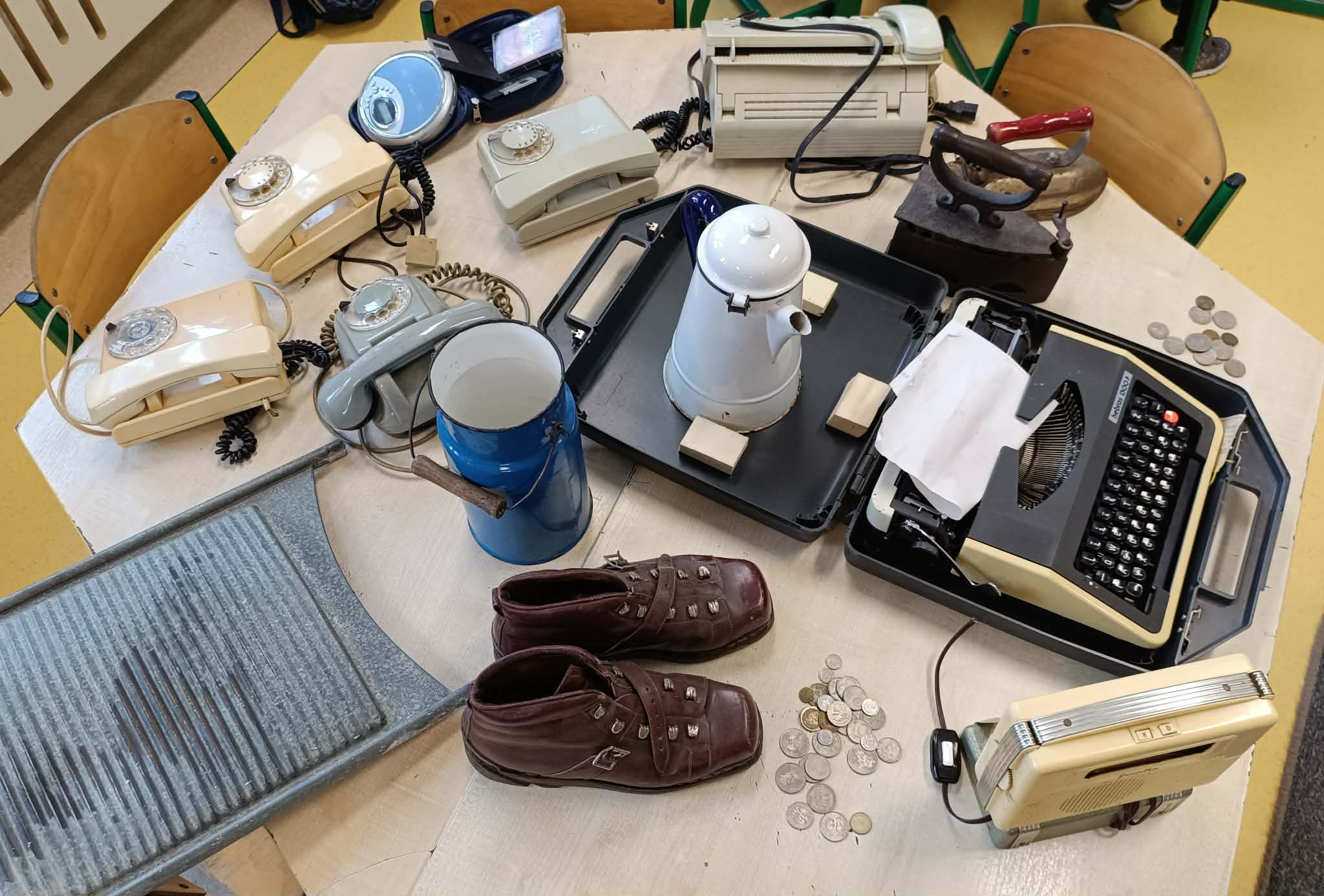 Na stole w sali leżą różne stare przedmioty codziennego użytku: kilka telefonów z tarczą, maszyna do pisania w walizce, metalowa tarka do prania, żelazko z duszą, dzbanek, radio, monety oraz buty narciarskie.