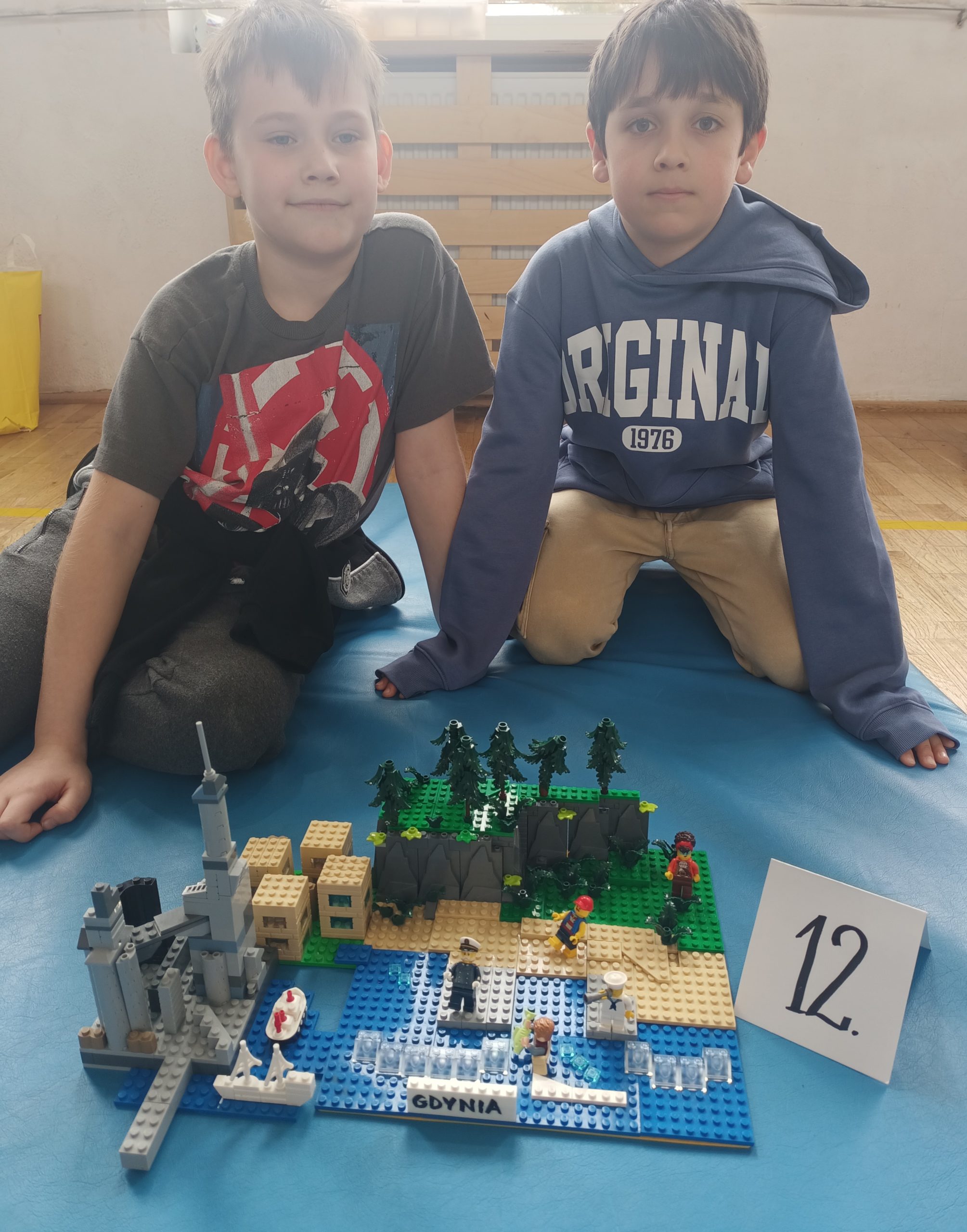 W sali gimnastycznej na niebieskim materacu siedzi dwoje uczniów z klasy 1. Przed nimi stoi ich budowla z klocków LEGO i etykietka z literą D.