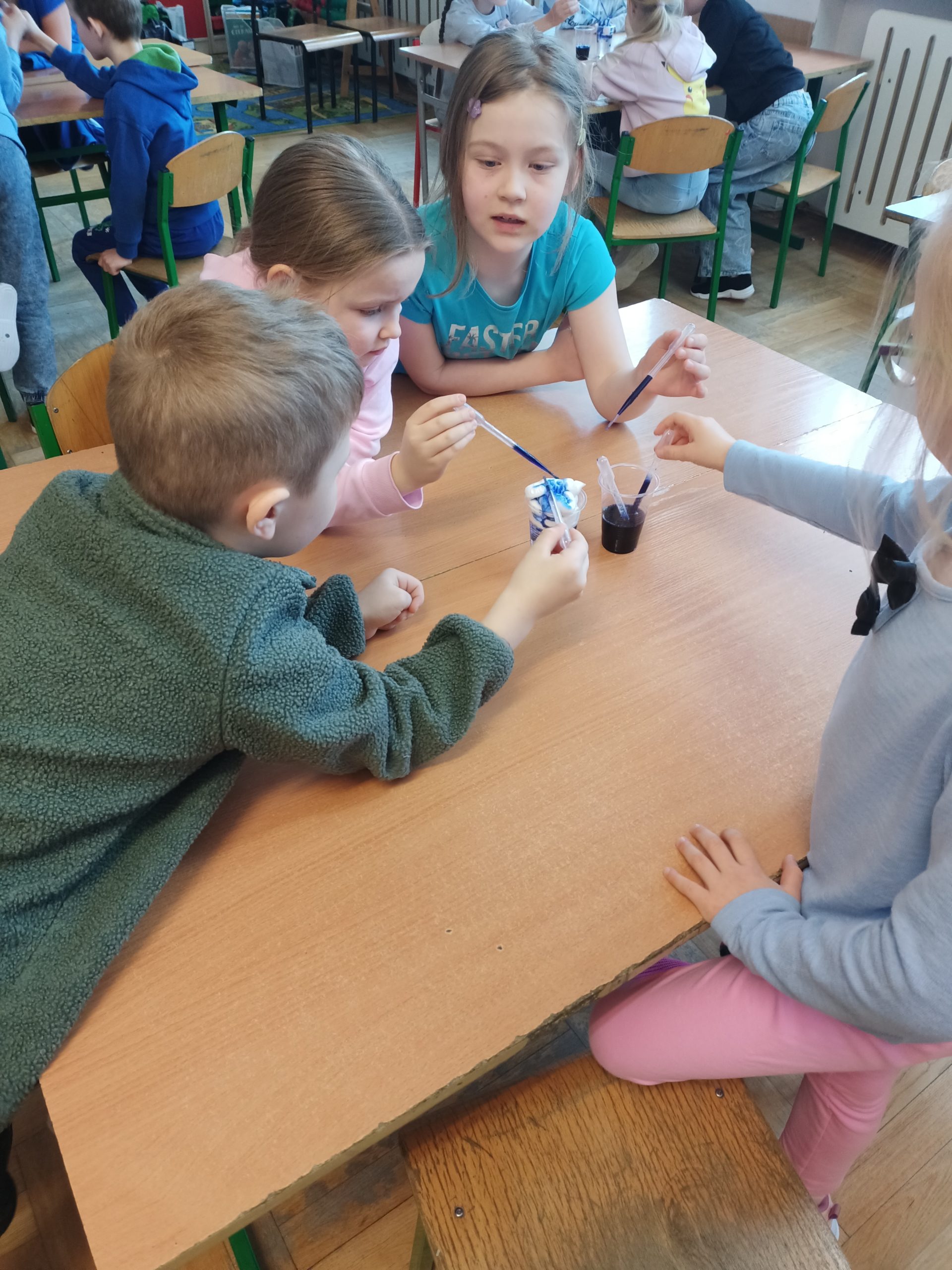 .Trzy dziewczynki i chłopiec siedzą w sali lekcyjnej po obu stronach ławki. N środku stoją dwa kubki z białą i niebieską cieczą. Dzieci używając kroplomierzy mieszają ciecze.